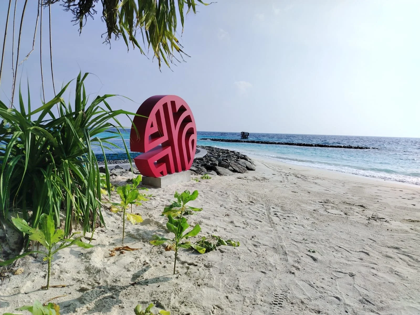 A sandy beach with a pink, maze-like sculpture near some green plants, a calm ocean in the background, and a partly cloudy sky.