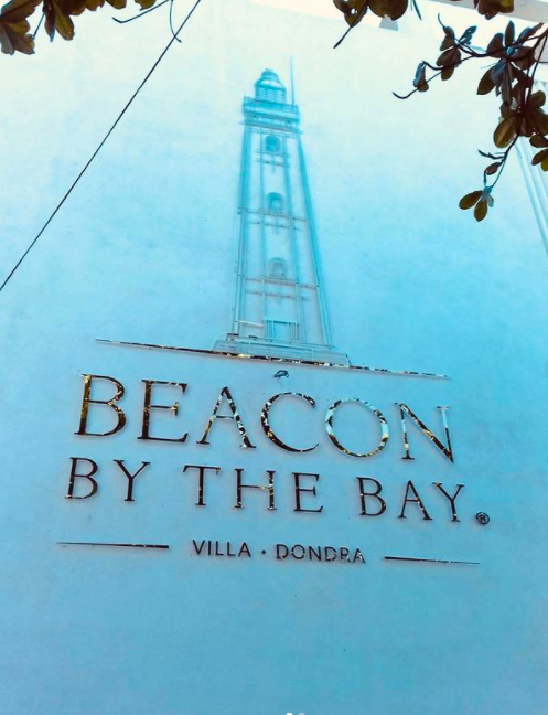 A sign reading "BEACON BY THE BAY VILLA DONDRA" with a tall lighthouse illustration above, and some leaves and wires in the foreground.