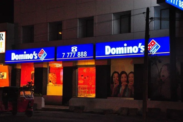 Exterior of a Domino's pizza restaurant at night with illuminated blue signage displaying the Domino's logo and phone number.