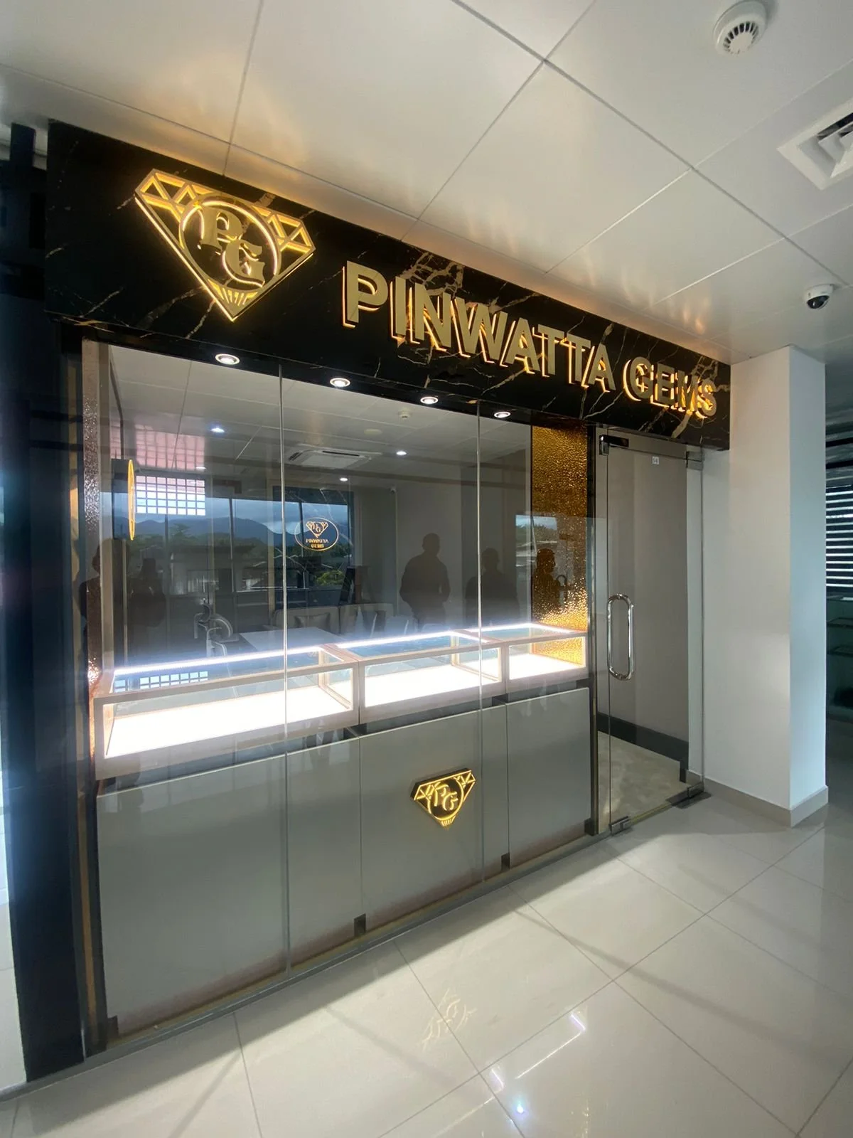 A jewelry store named Pinwatta Gems with a black marble sign, gold lettering, and a diamond-shaped logo. The store has glass display cases, and there are reflections of people and the surrounding area visible on the glass.