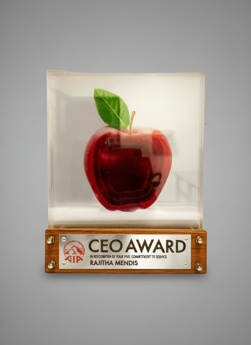 Red apple with green leaf inside a clear display case, mounted on a wooden plaque labeled "CEO AWARD" with the recipient's name Rajtha Mendis.