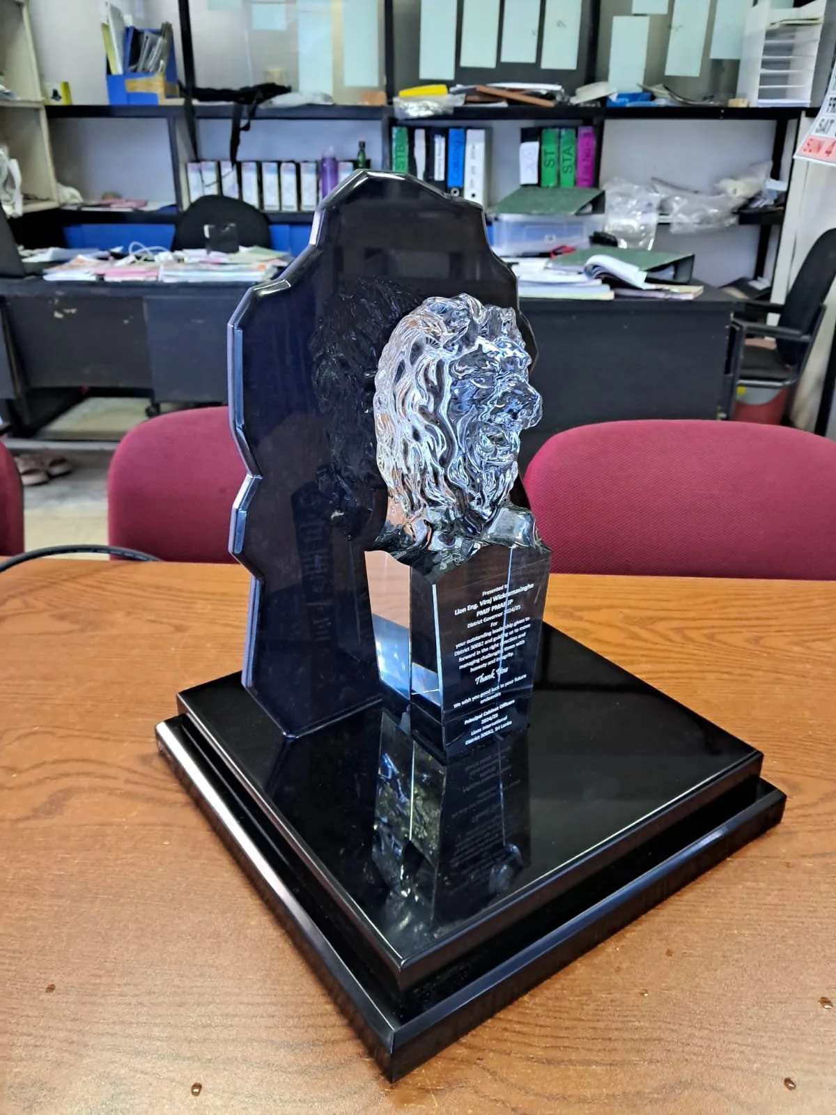A trophy featuring a profile of a lion's head mounted on a black base, placed on a wooden table in an office setting with shelves, binders, and papers in the background.