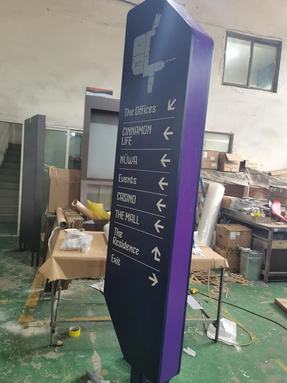 A tall, vertical directional sign in a warehouse or workshop. The sign has blue and purple colors with white text and arrows, pointing to various locations such as The Offices, Cinnamon Life, Nüwa, Events, Casino, The Mall, The Residence, and Exit.