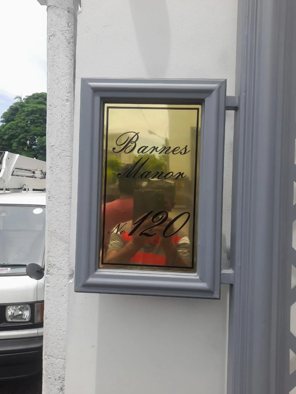 Reflective glass sign with black cursive text reading "Bannes Manor No 120" in a gray frame on an exterior wall, with a person taking the photo visible in the reflection.