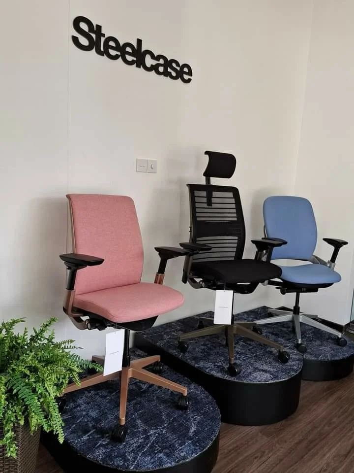Display of three office chairs in front of a white wall with the 'Steelcase' logo, arranged on black circular platforms, with a green plant to the side.