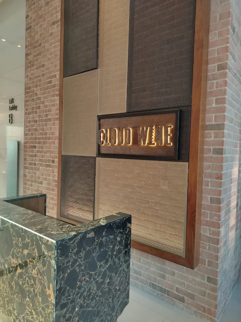 Interior view of a modern lobby with a sign that reads "CLOUD WINE" mounted on a wall with a dark and beige textured panel background, brick wall, and a black marble reception desk.
