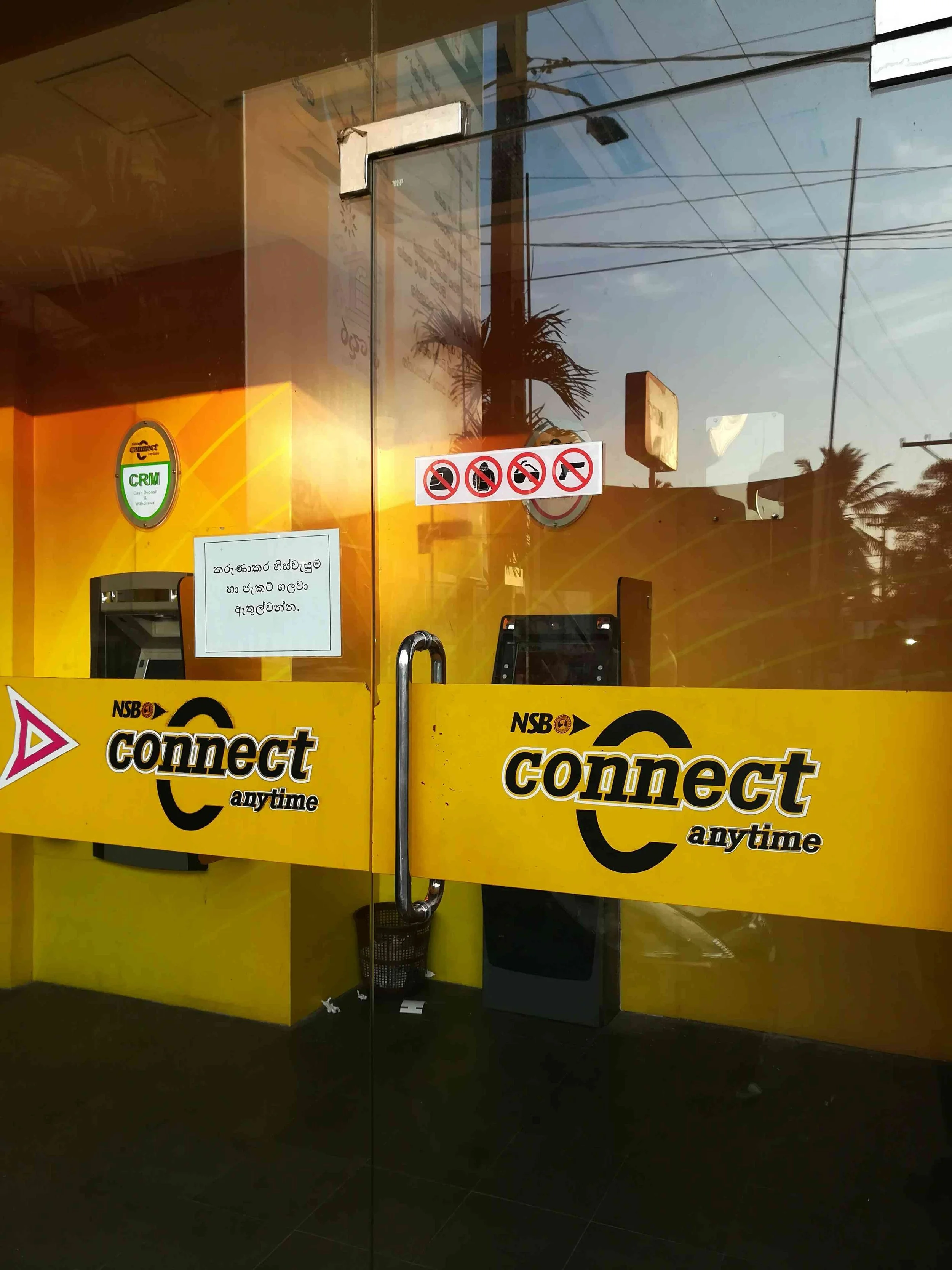 Glass door with yellow and black signage reading 'connect anytime,' a sign showing no smoking, no cameras, no pets, no food or drinks, and a palm tree reflection outside.