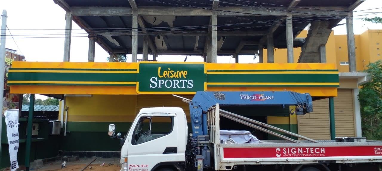 An unfinished two-story building with a green and yellow sign that reads "Leisure SPORTS" on the ground level. A blue cargo crane truck is parked in front of the building, partially blocking the view.
