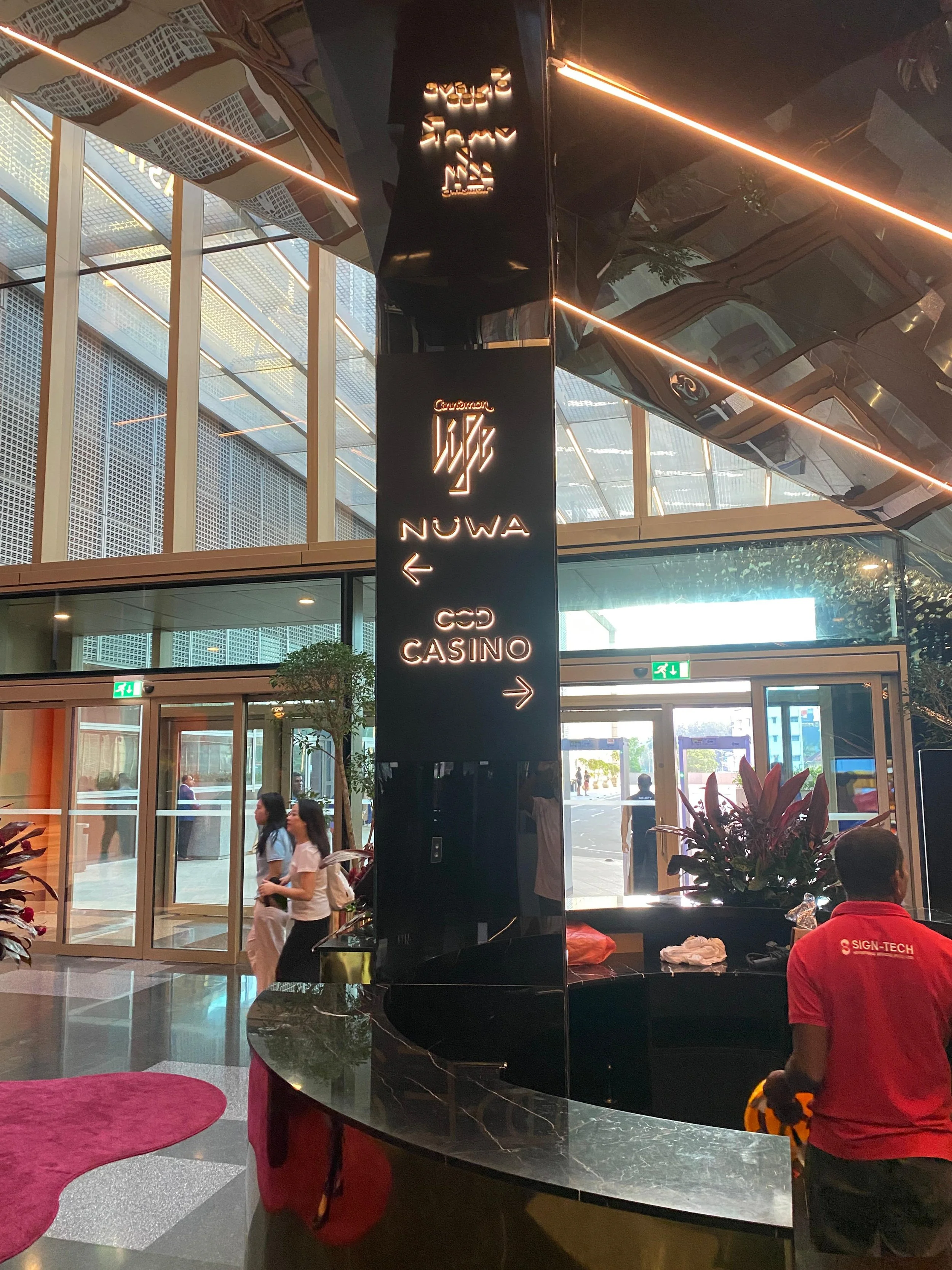 Interior of a modern casino or entertainment complex with glass walls, people walking, and a black signpost with directional signs to a casino and other areas.