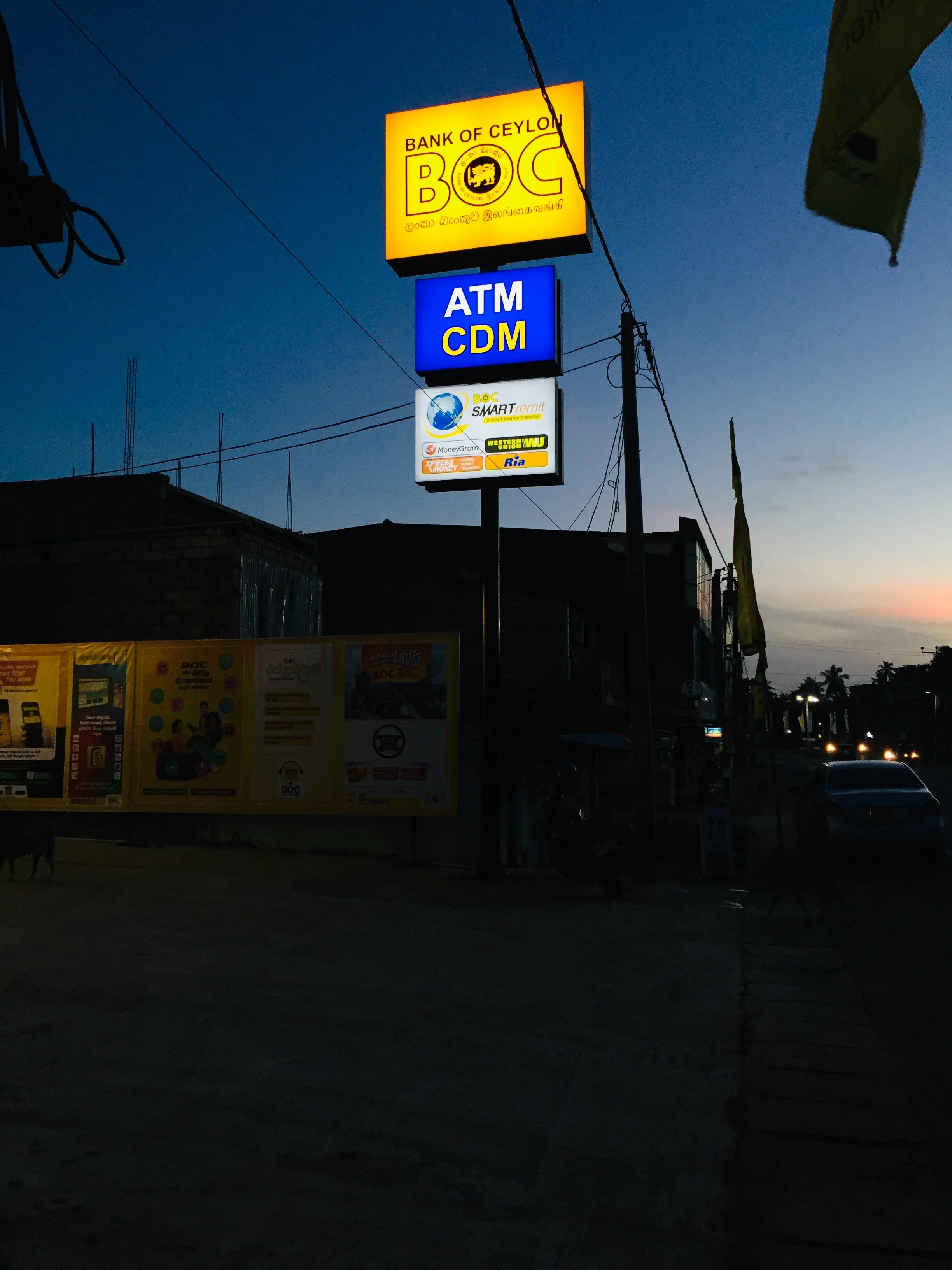 Signboard at dusk showing the Bank of Ceylon logo and name, along with signs for ATM and CDM services, and other financial service advertisements.