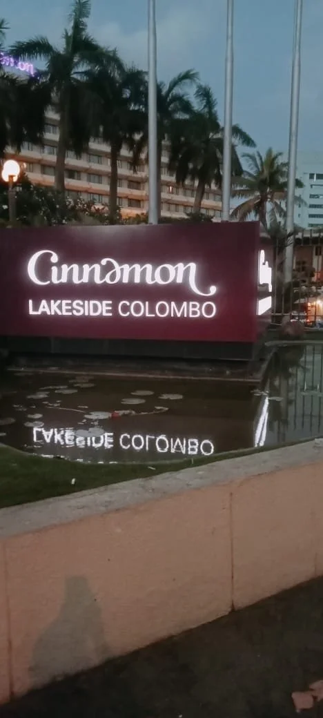Sign for Cunndmon Lakeside Colombo with palm trees and buildings in the background during evening.