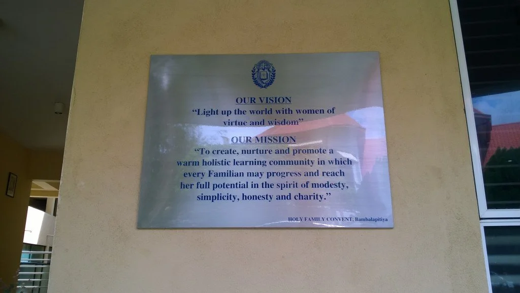 A metallic plaque displaying the vision and mission statement of Holy Family Convent, Bambalapitiya, mounted on a yellow wall.