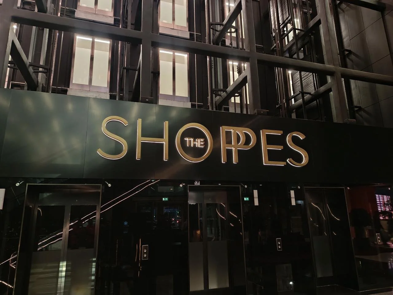 Sign for The Shops at night with illuminated letters on a dark building facade.