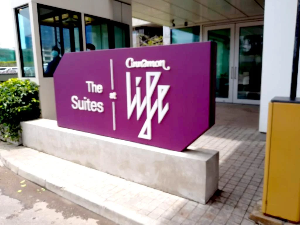 A purple sign outside a building reads 'The Suites at Guzman.' The building has glass doors and windows, with a small bush on one side and a yellow box on the other.