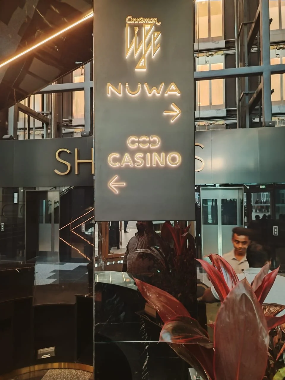 Indoor location with illuminated signs directing to NUWA, CCD Casino, and an arrow pointing right. Reflections of people and plants are visible on glass surfaces.