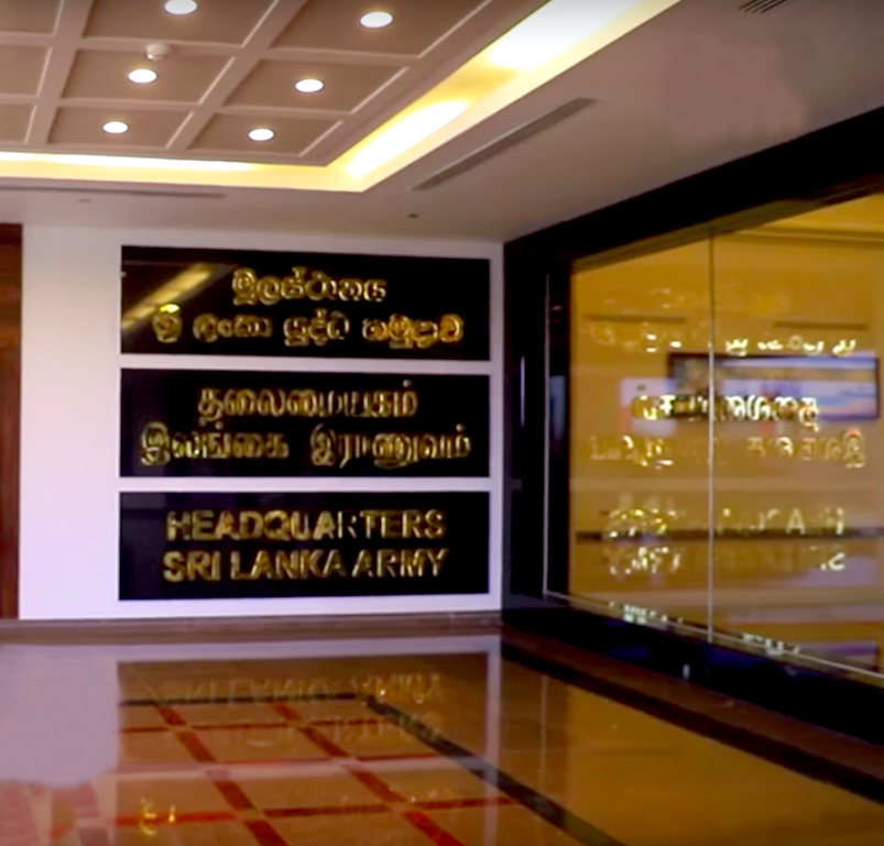 Sign in Sri Lanka Army headquarters with text in three languages, including English, on a black background with golden lettering, near a glass wall in an indoor setting.