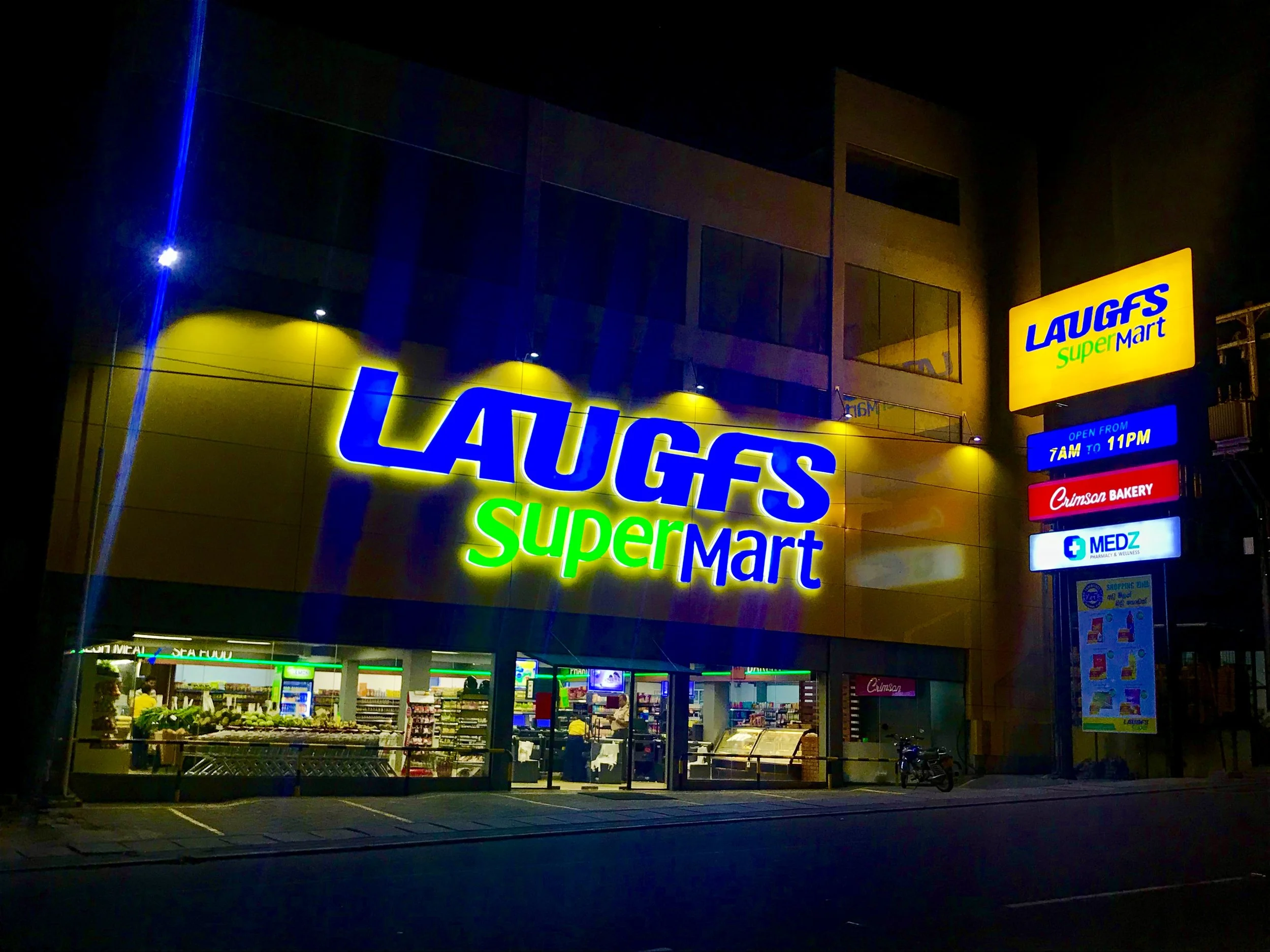 Nighttime view of a Laugfs Supermarket with brightly lit yellow and blue signage. The store's interior is visible through the glass entrance, with illuminated shelves and shopping carts outside.