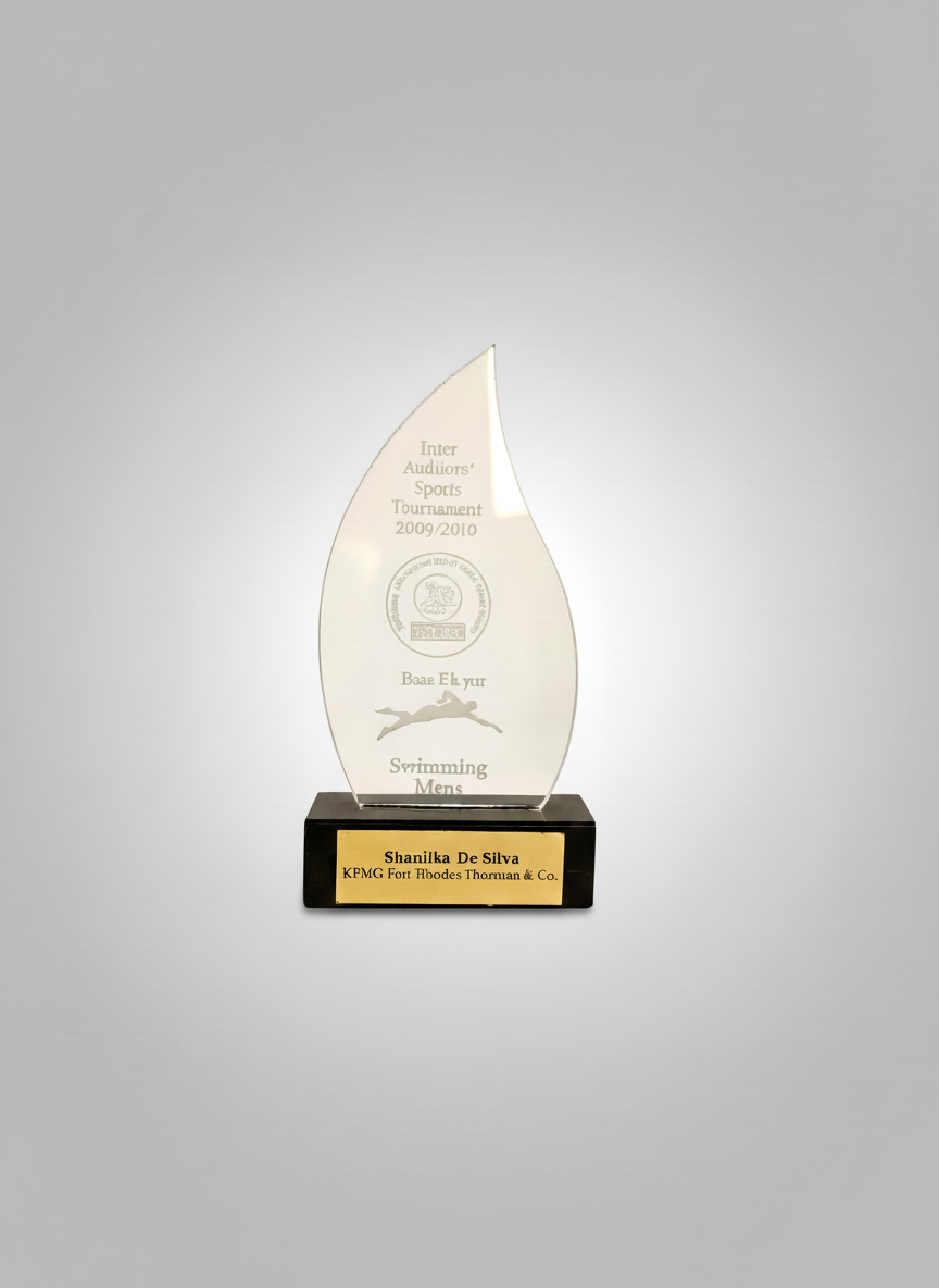 Glass award sculpture with a black base and gold plaque, engraved with the name Shanilka De Silva and KPMG Fort Bhodes Thormian & Co. The sculpture has text about a 2009/2010 inter-audiators' sports tournament, featuring a logo, and the words 'Bae Eh