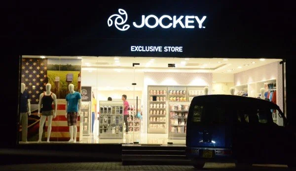 Front view of a Jockey store at night, illuminated, with mannequins displaying clothing and accessories inside, and a van parked outside.