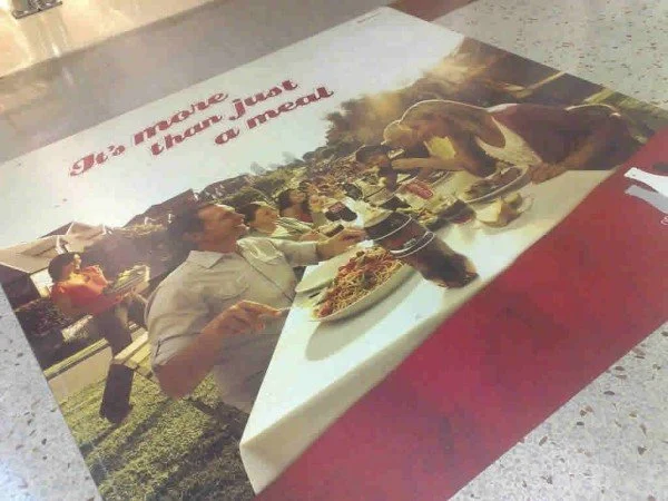 A restaurant table with a printed photo on the tablecloth showing people eating pizza and socializing outdoors under a tent. The photo has the text 'Let's make you know just a meal' printed at the top.