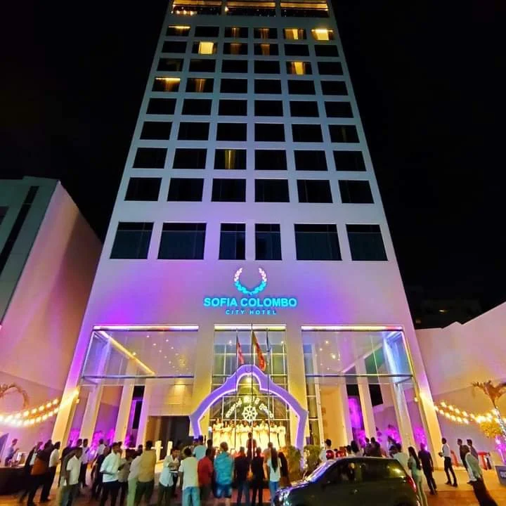 Nighttime view of the Sofia Colombo City Hotel with purple and yellow lighting, featuring a crowd of people outside and the hotel entrance decorated at the front.