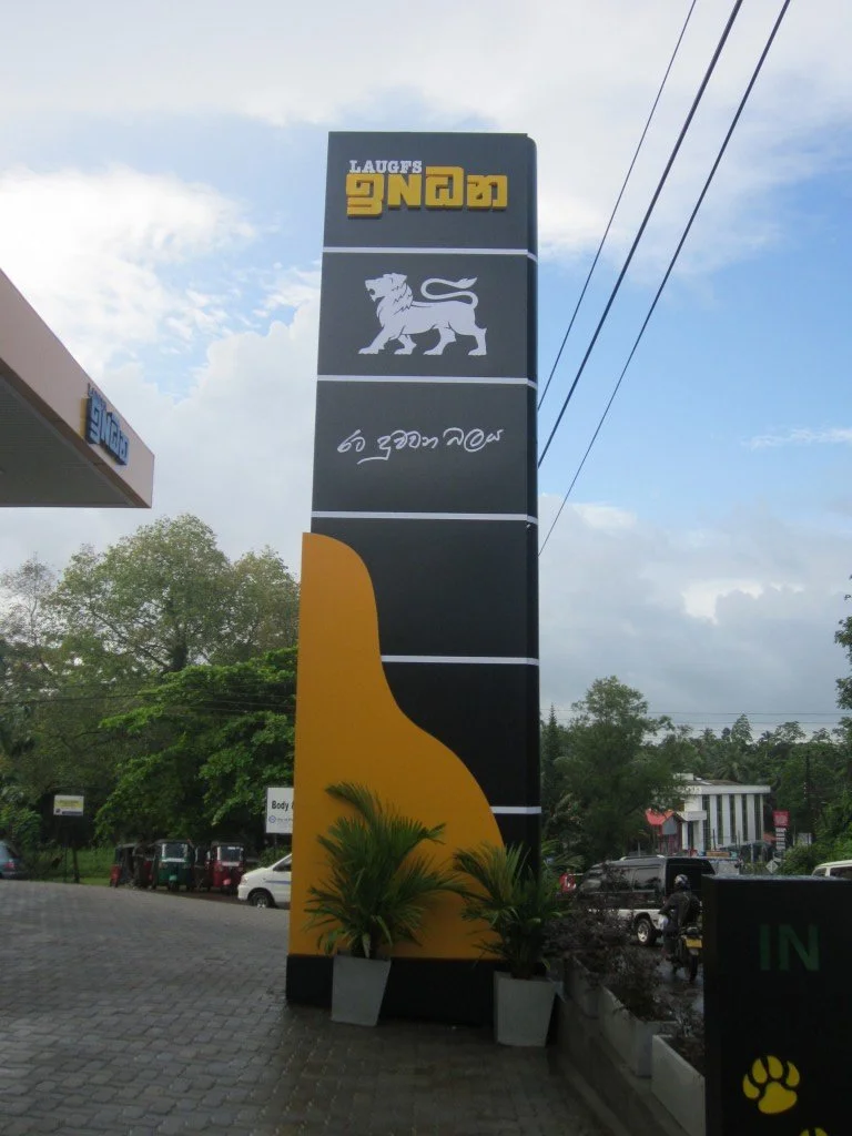 Tall sign with black, yellow, and white colors displaying a lion's head logo and text in a foreign script, outside in a parking lot with trees and buildings nearby.