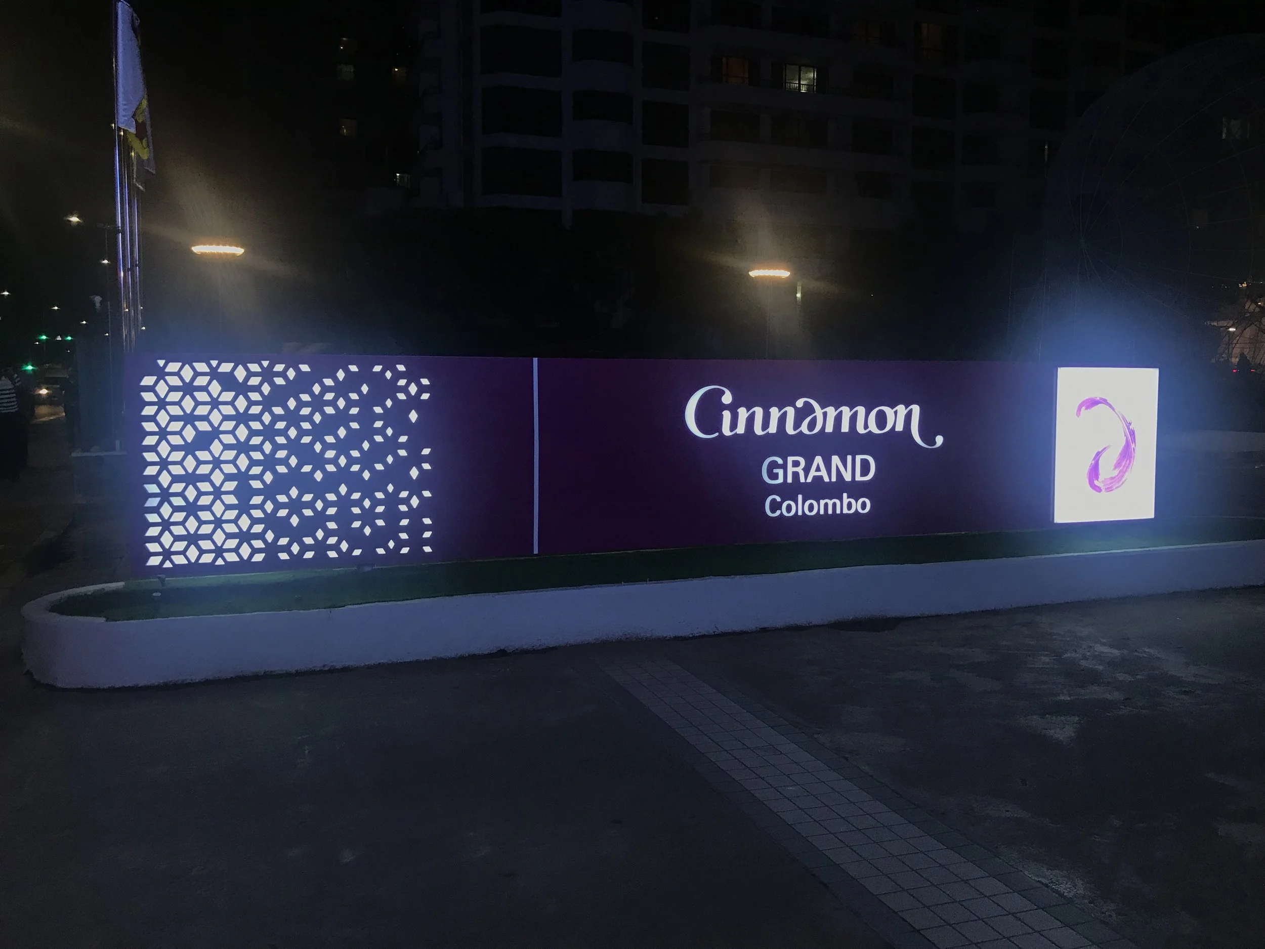 A brightly lit promotional sign at night displays for an event called 'Cinnamon Grand Colombo.' The sign features a purple background with white geometric patterns and text, and an image of a purple swan on the right side.
