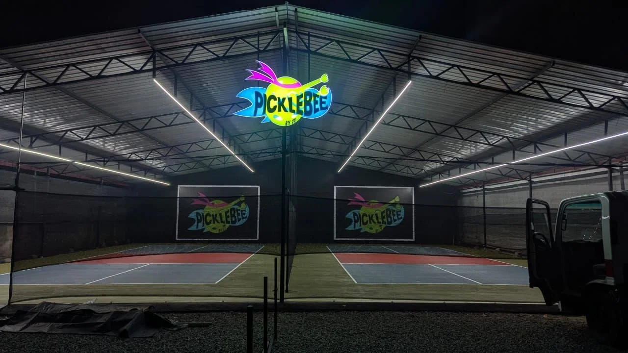 Night view of an illuminated outdoor pickleball court with a colorful Picklebee sign hanging above.