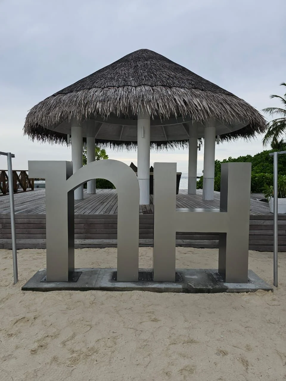 A metallic sculpture of the letters 'PH' in front of a thatched-roof pavilion on a sandy beach.