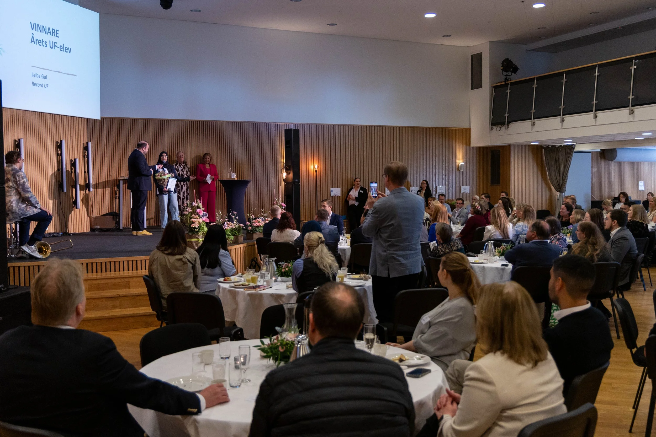 En prisutställning eller ceremoni med en person som tar emot ett pris på scenen, där en stor skärm visar texten 'VINNARE Årets UF-elev Laiba Gul Record UF'. En publik är samlad och några personer tar bilder.