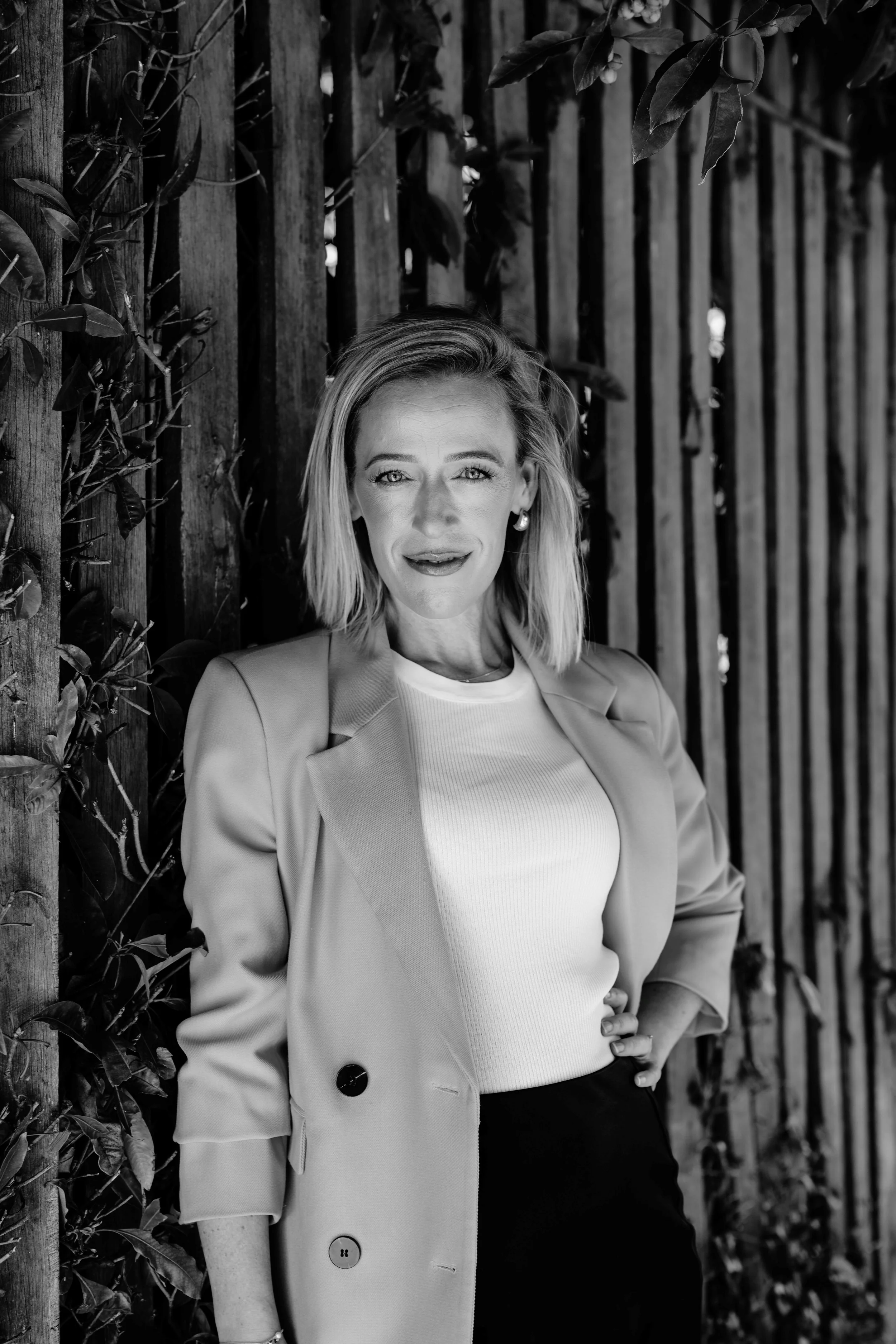 A woman with shoulder-length hair, wearing a blazer over a white top, stands outdoors against a wooden fence with foliage, smiling at the camera.