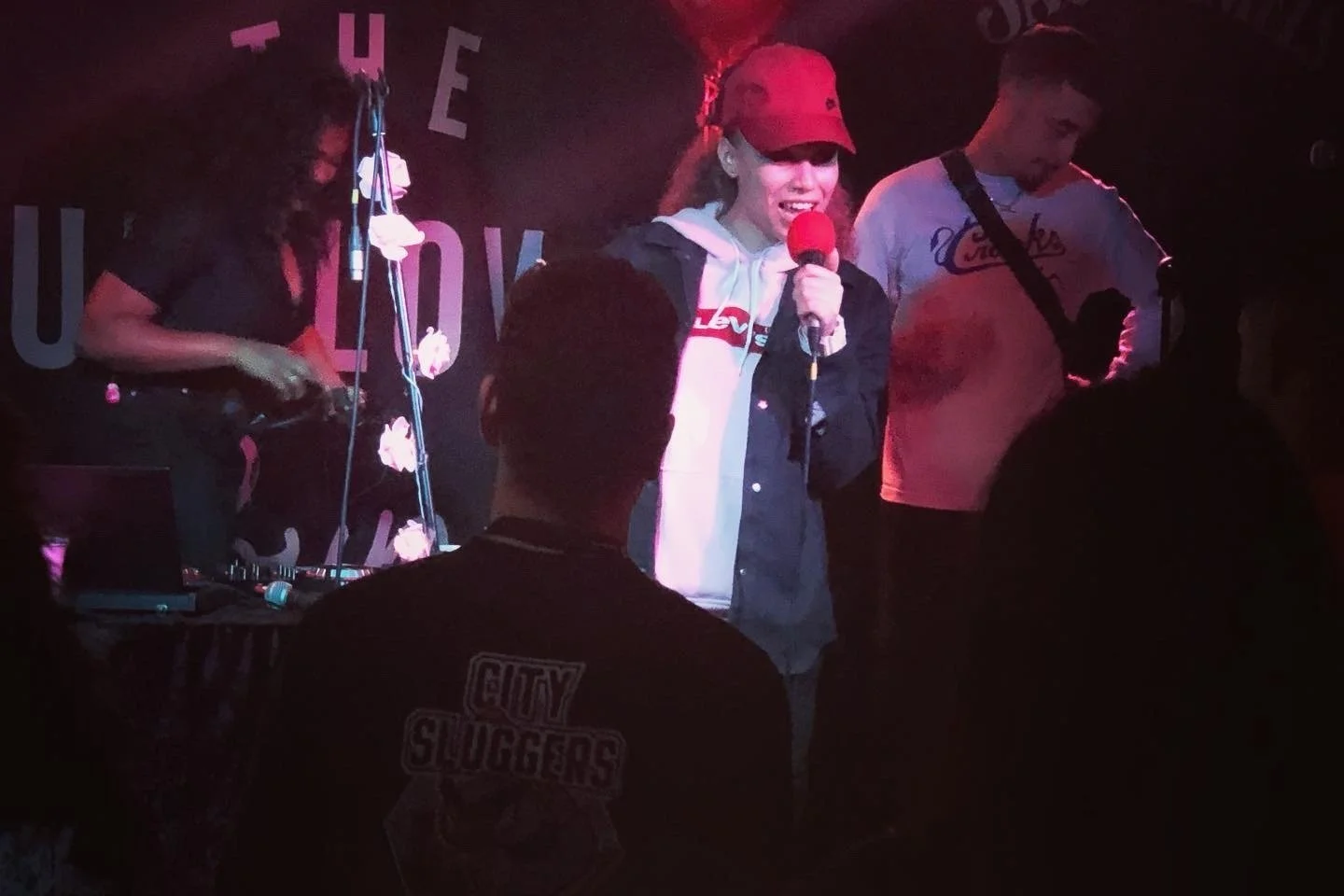 A woman holding a red microphone performing on stage with two men, one using DJ equipment, at a nightclub or music venue.