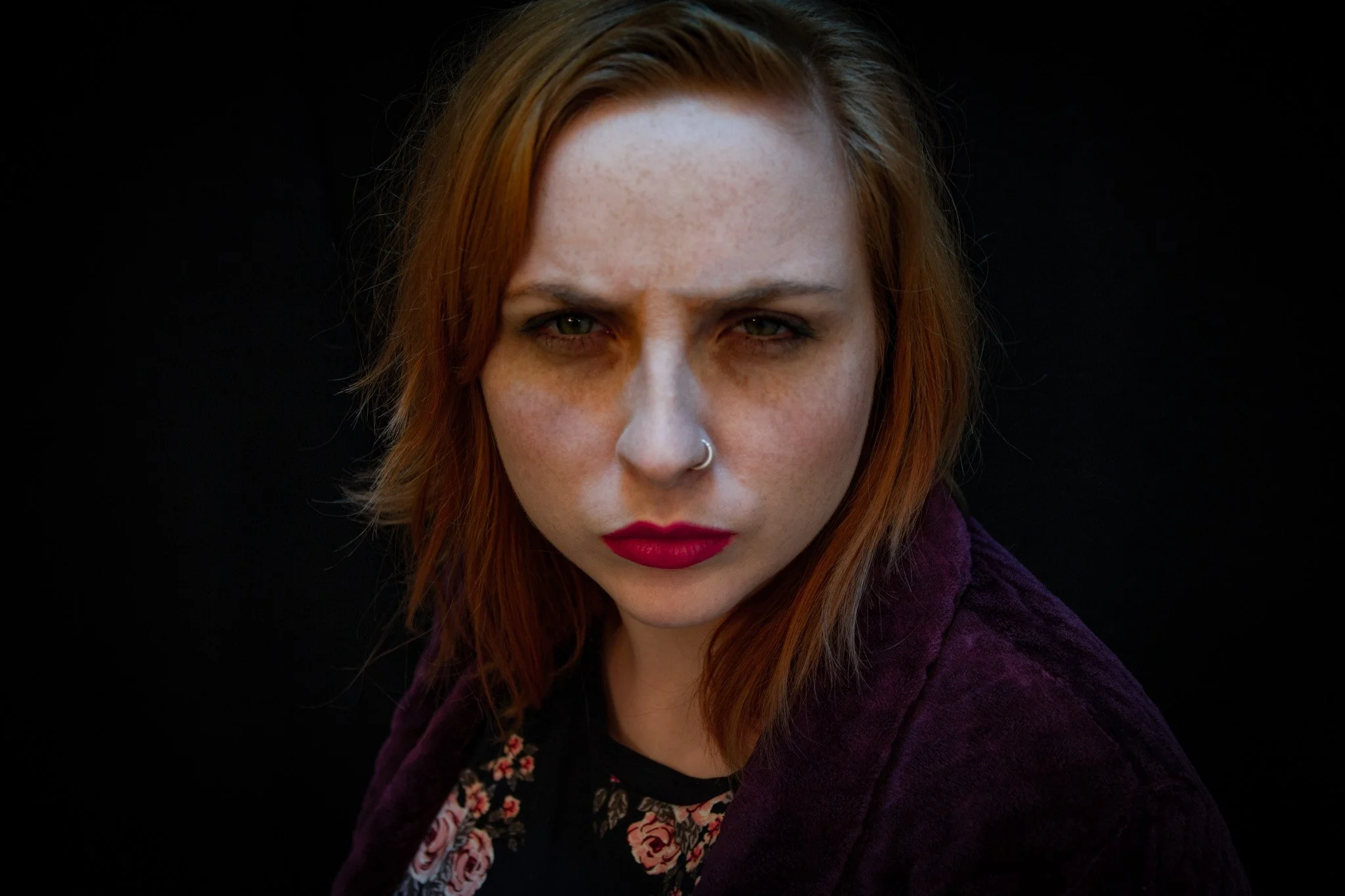 Close-up of a woman with reddish-brown hair, green eyes, and a nose piercing, wearing a dark top and a purple jacket against a black background.