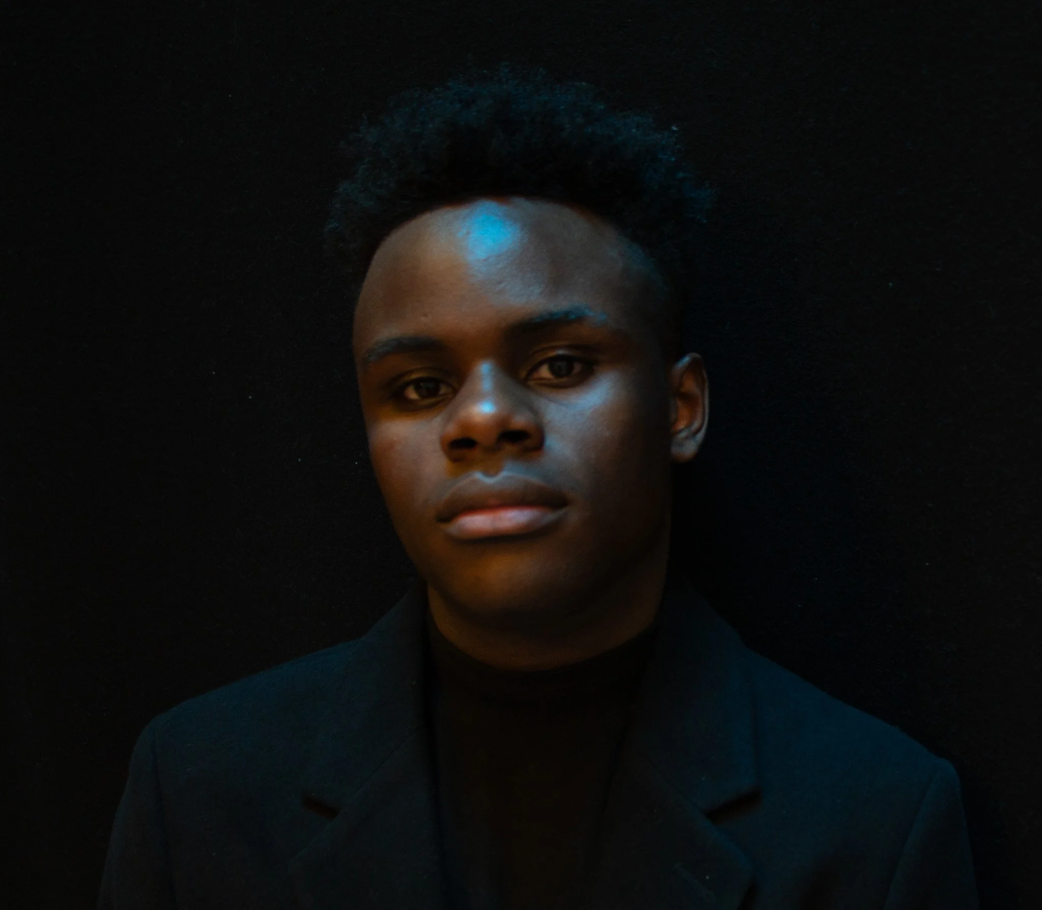 Portrait of a young man with dark skin, wearing a black blazer, against a dark background, looking directly at the camera.