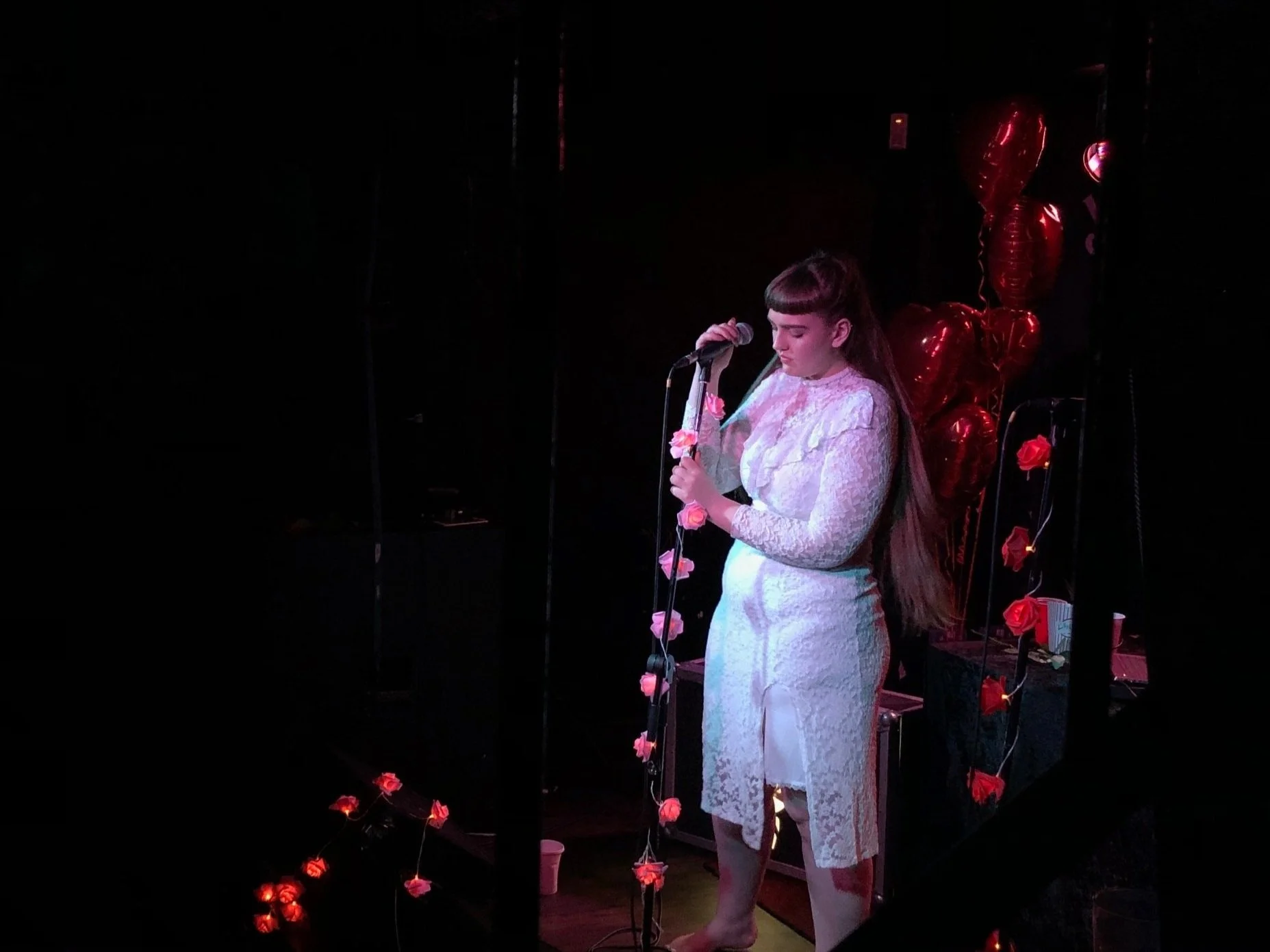 A woman in a white lace dress standing on stage, holding a microphone, with red heart-shaped balloons and string lights decorated with pink roses behind her, in a dark setting.