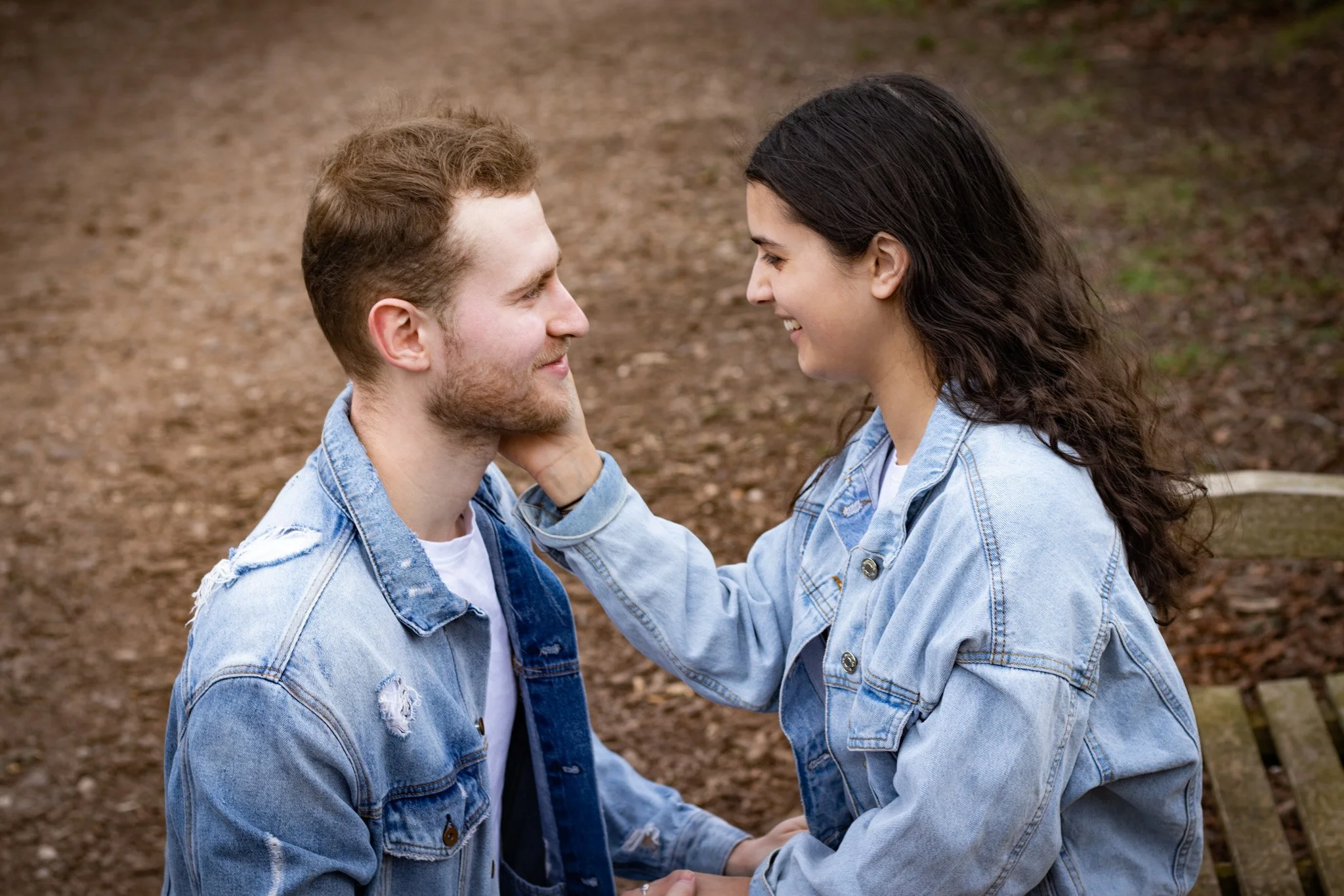 A young man and woman sit close together outdoors, smiling and looking into each other's eyes. The woman gently touches the man's face. They are both wearing light denim jackets, with a blurred natural background.