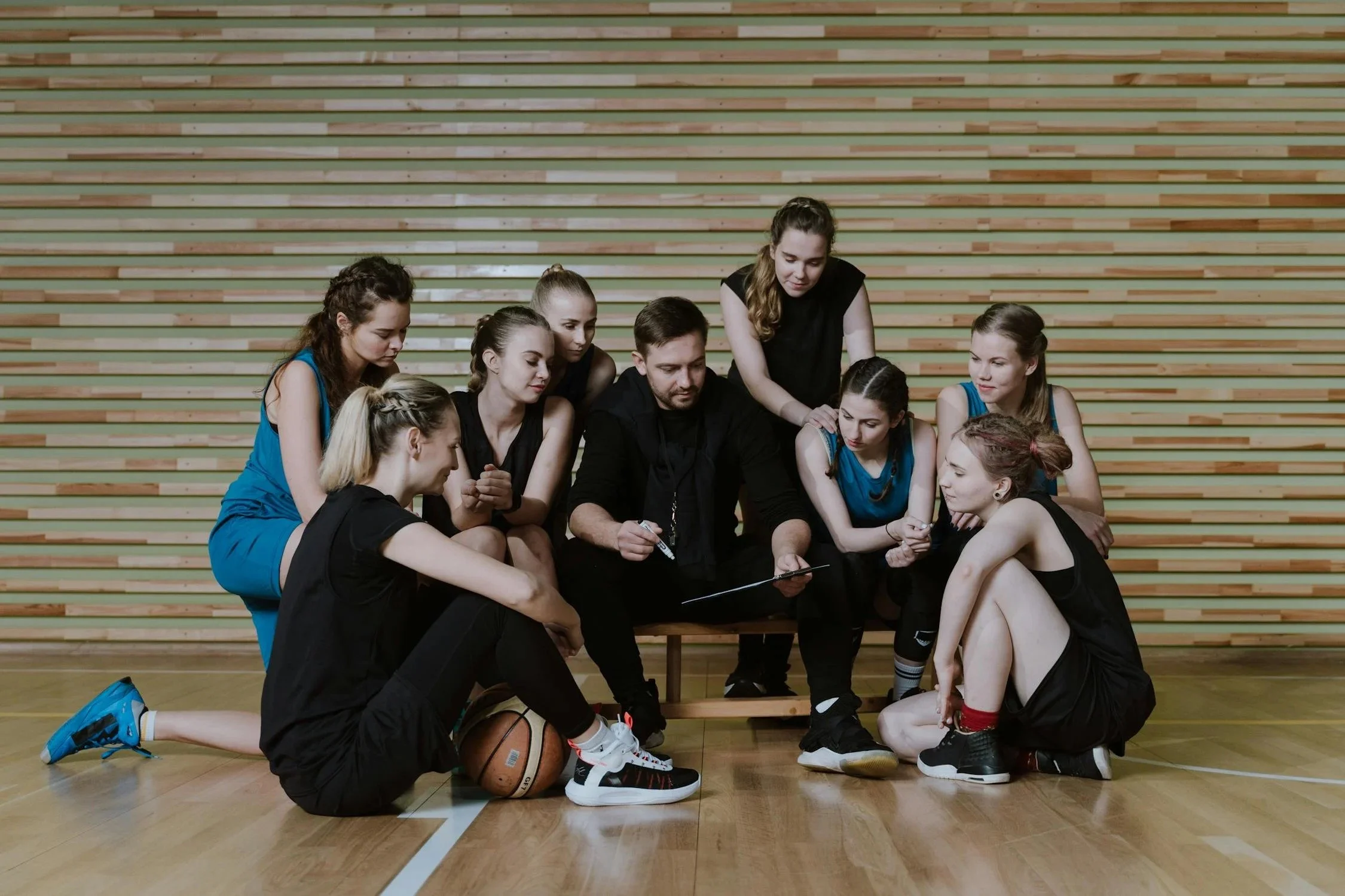 Basketball coach and team girls gathered on a basketball court, listening to instructions with a basketball on the floor.