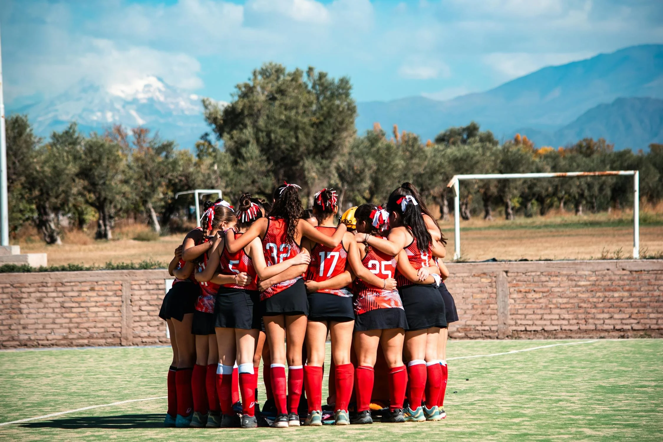 A group of young female rugby players in red and black uniforms huddling together on a field with mountains and trees in the background.
