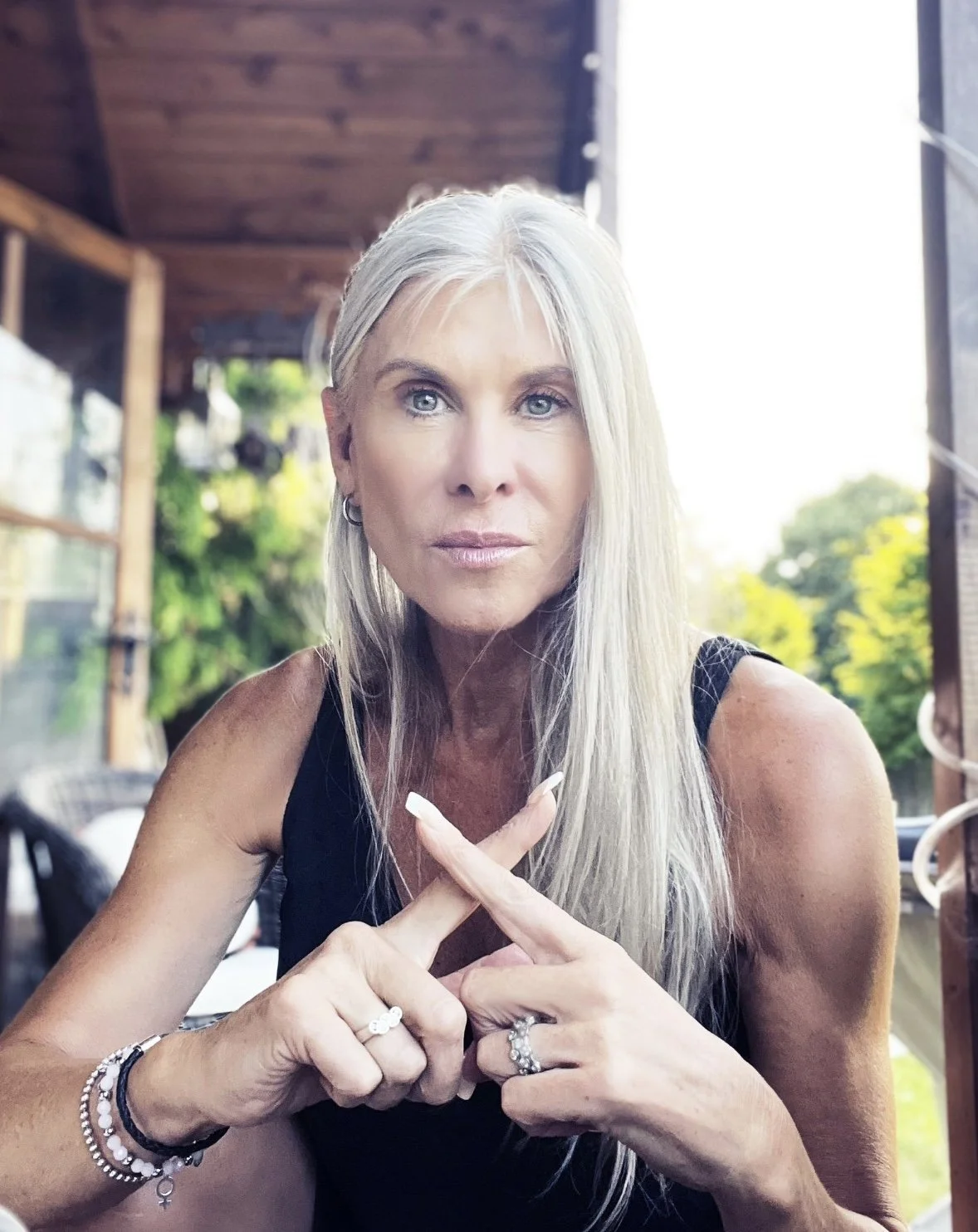 A woman with long, straight, gray hair and blue eyes looking directly at the camera, making an 'X' shape with her hands, seated outdoors with trees and a wooden structure in the background.