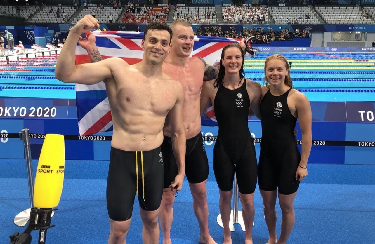 Four Olympic swimmers, two men and two women, standing side by side on a swimming pool deck, celebrating and smiling; two of the athletes are holding a Union Jack flag behind them, with