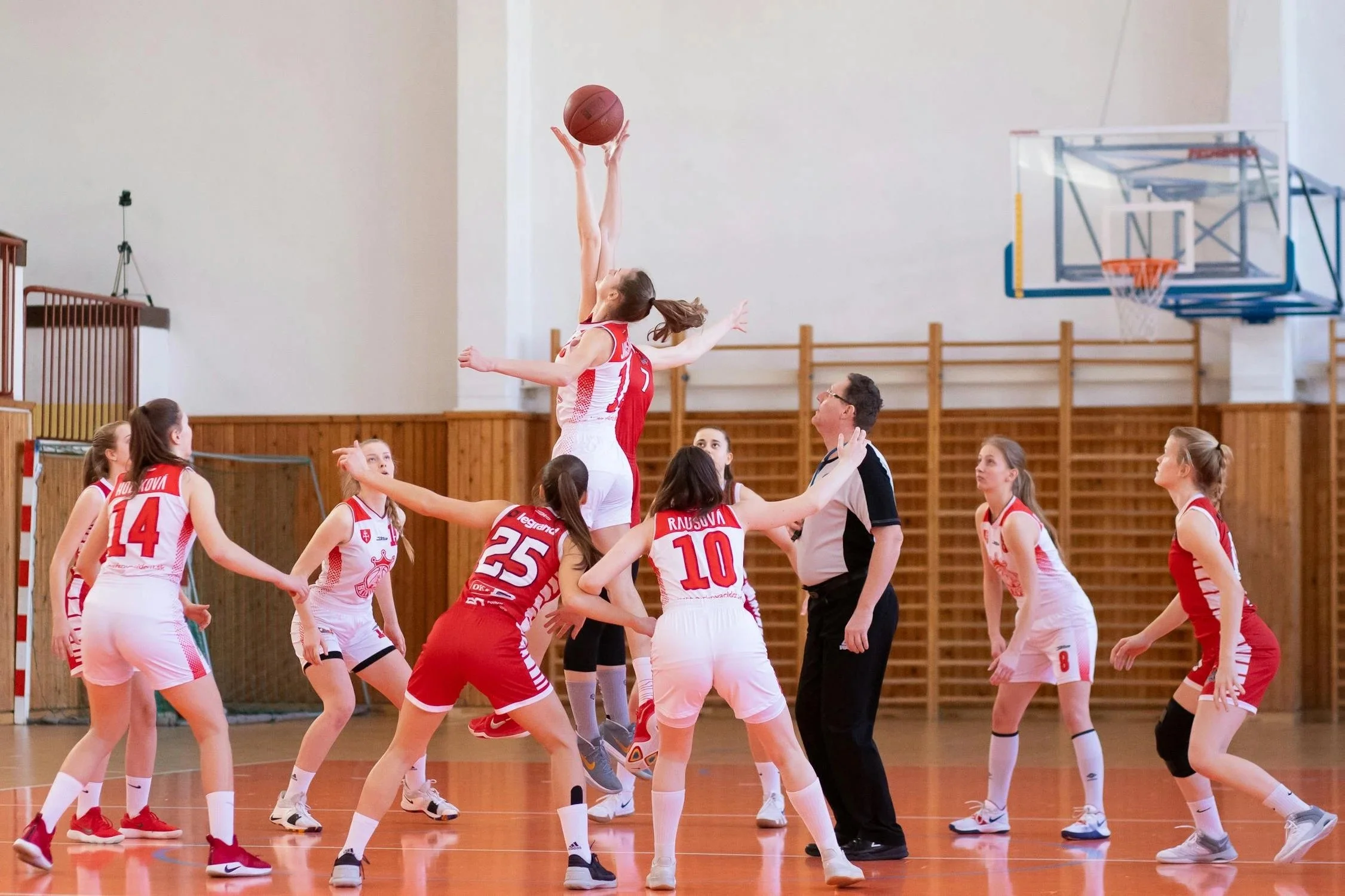 A girls' basketball game in progress, with a player in a white uniform jumping to shoot the ball while other players and a referee look on inside a gymnasium.