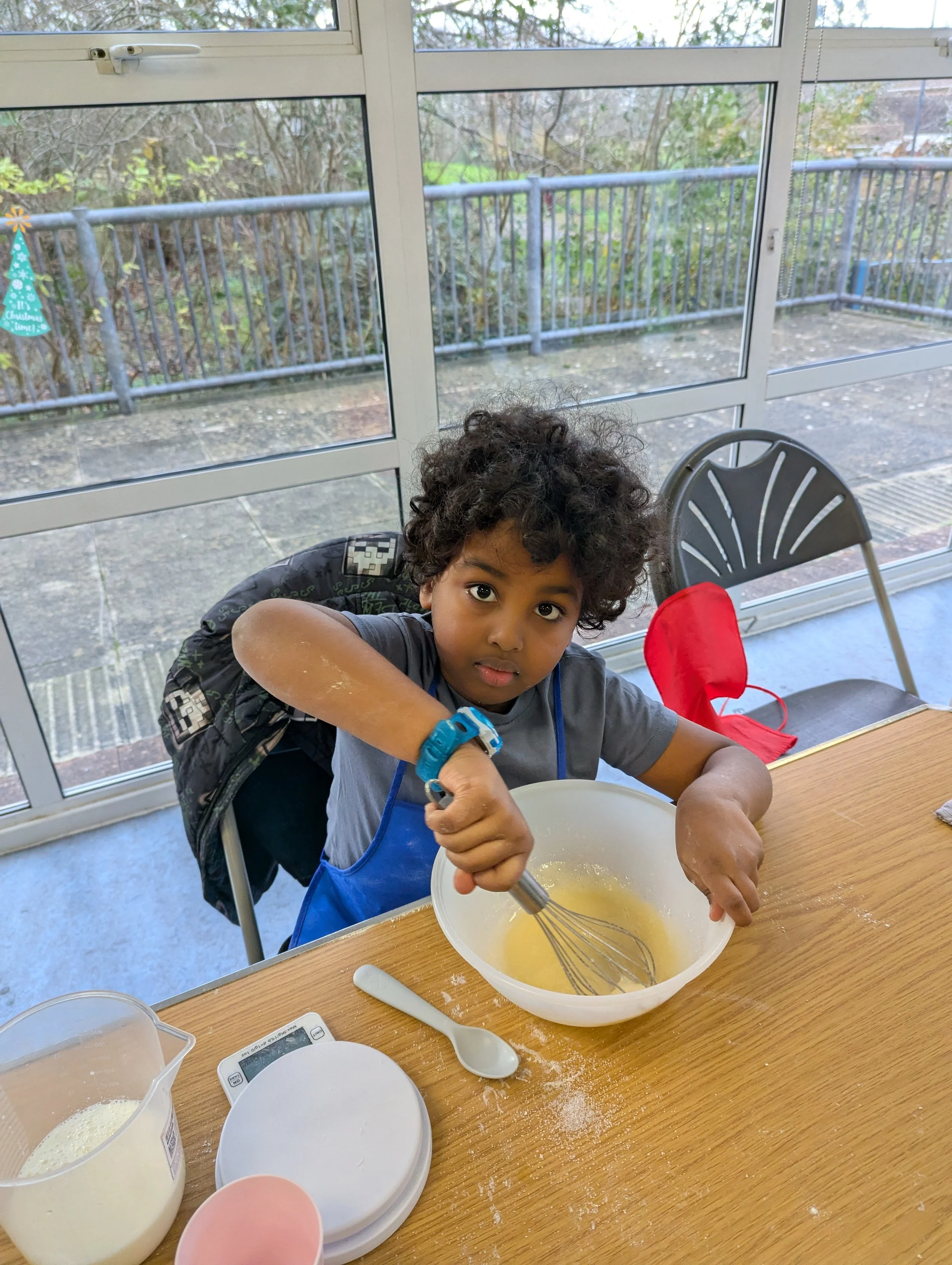A young child with curly hair, wearing a gray shirt and a blue apron, is mixing batter in a bowl with a whisk at a wooden table. There are baking ingredients, a spoon, and a digital scale on the table. The child is sitting in a room with large windows showing an outdoor balcony or patio area.
