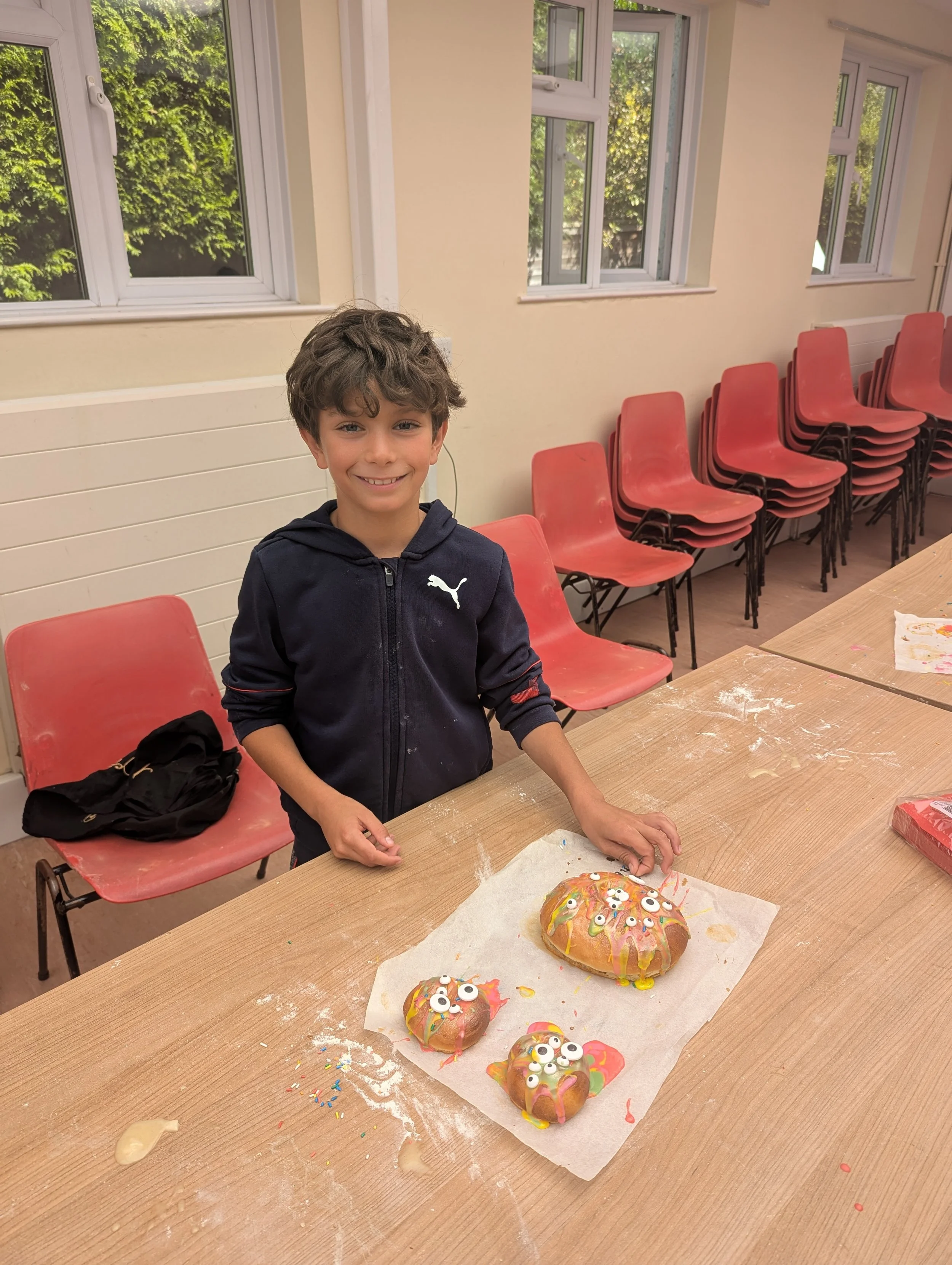 A young boy with curly hair smiling at the camera, standing next to decorated decorated baked treats that look like Halloween monster heads with googly eyes, on a paper-lined wooden table in a room with red chairs and large windows showing greenery outside.