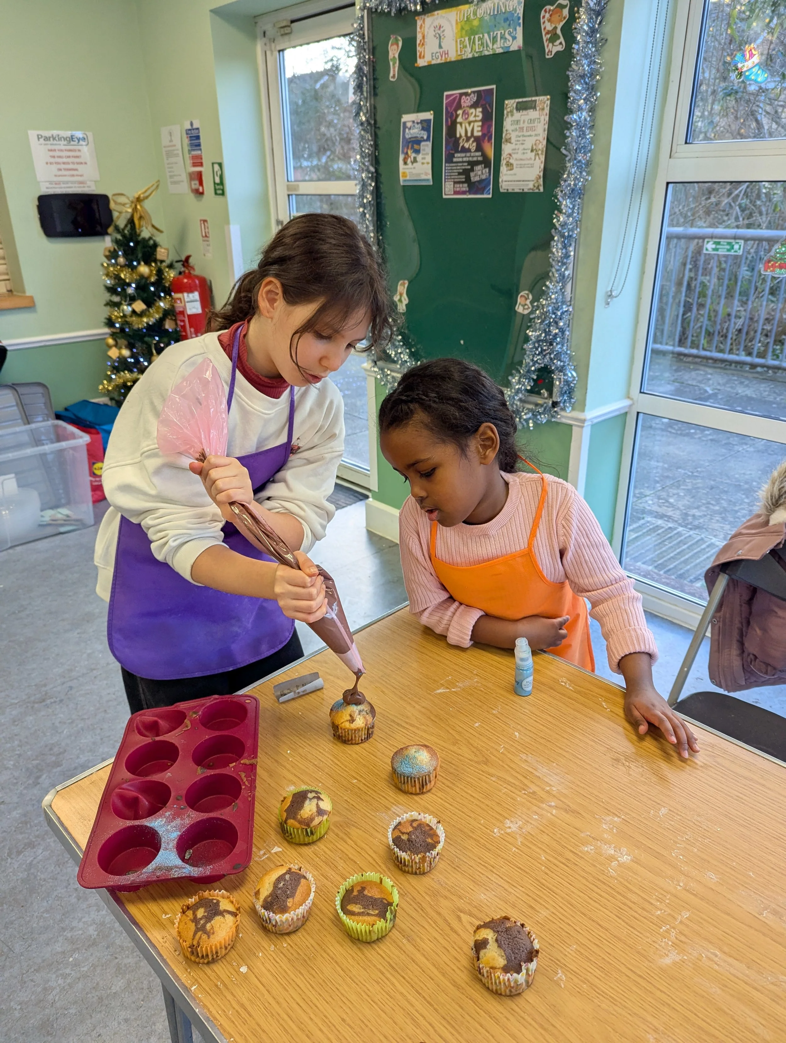 Two girls decorating cupcakes at a wooden table with a sprinkle of icing and topping. The girl on the left is using a piping bag with chocolate frosting, wearing a beige sweater and purple apron, and the girl on the right is watching, wearing a pink sweater and orange apron. There are uniced cupcakes on the table, a red cupcake mold, a small bottle of icing, and a festive Christmas tree in the background.