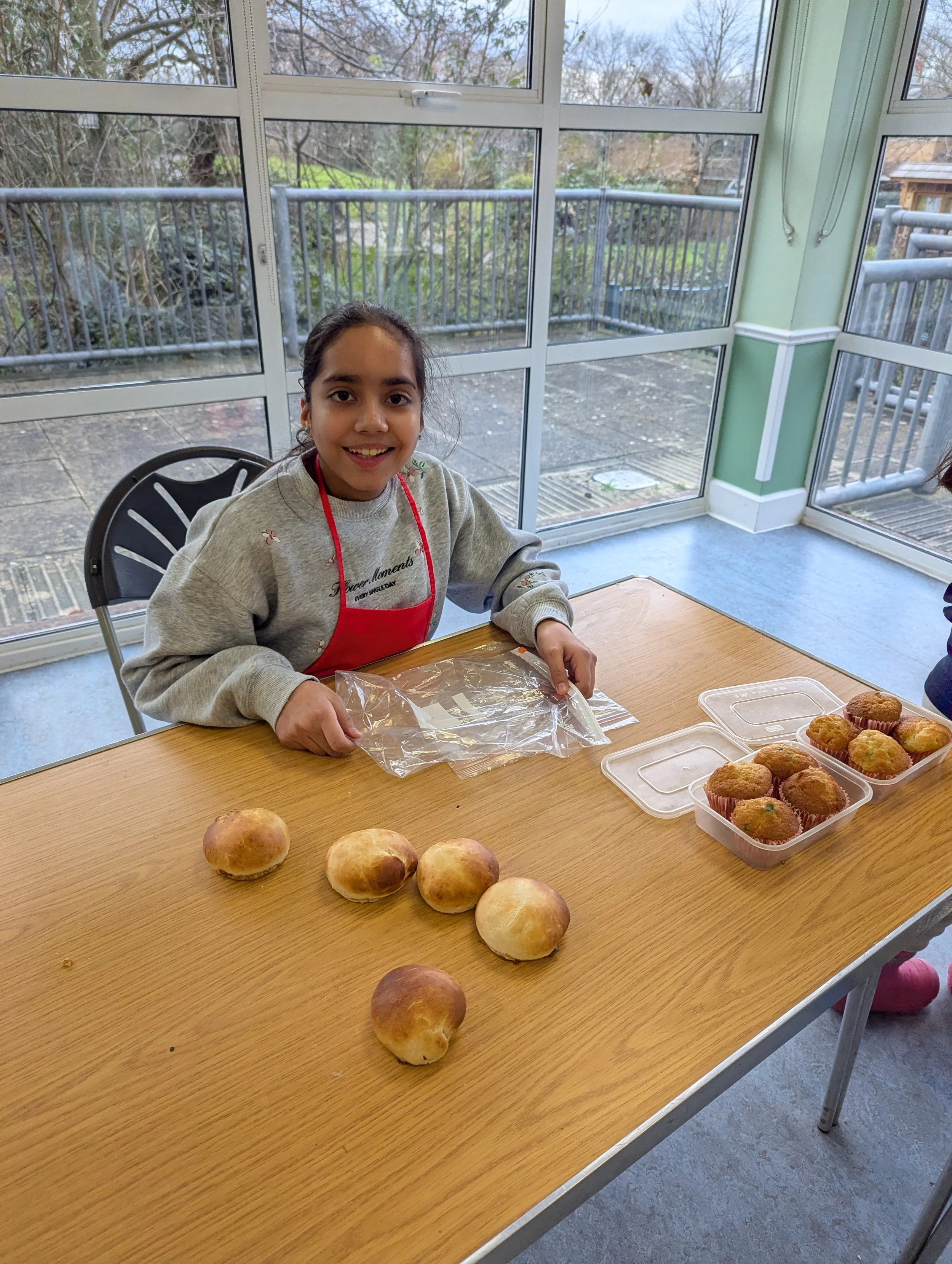 A young girl with dark hair in a ponytail, wearing a gray sweatshirt and red apron, sitting at a wooden table with baked goods including muffins and rolls, inside a bright room with glass windows and a balcony outside.