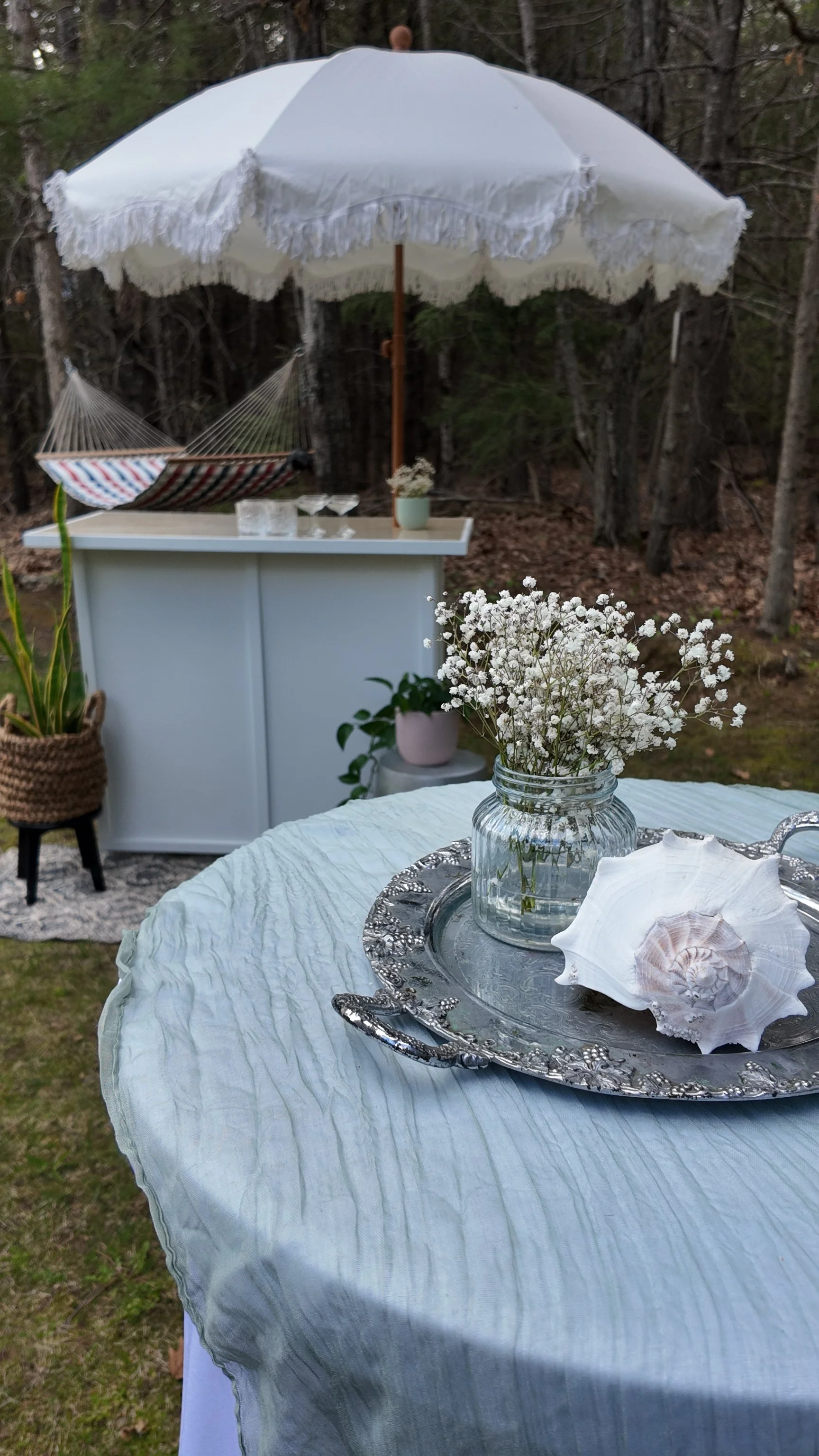 An outdoor setup with a round table covered with a light blue tablecloth, a silver tray with seashells, a glass jar with white flowers, and a decorative shell. Behind the table is a white counter with a potted plant, glasses, and a hammock under a large white umbrella with fringed edges. The background shows trees and a natural wooded setting.