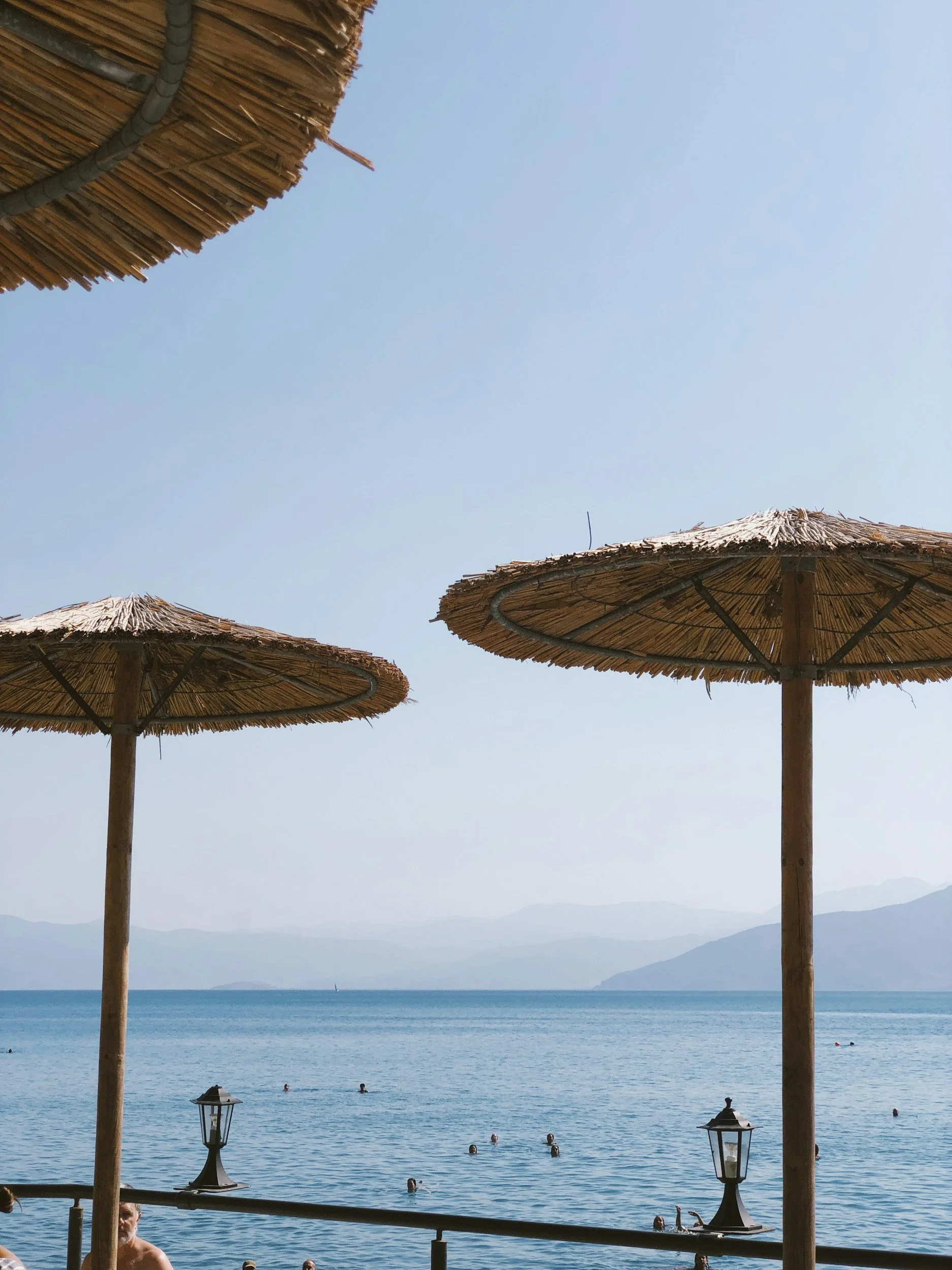 Beach scene with straw umbrellas, people swimming in the ocean, and a mountain in the background under a clear blue sky.