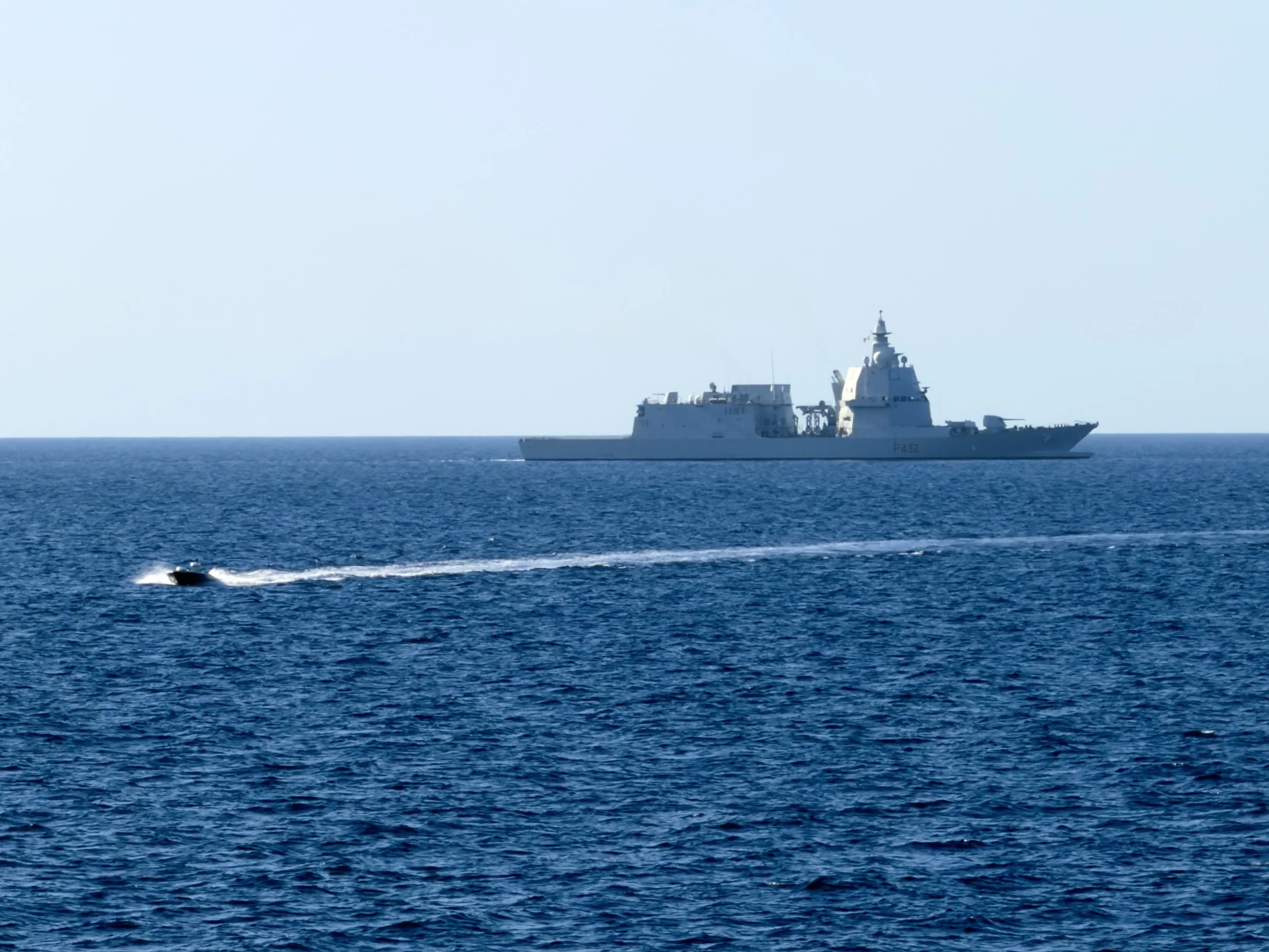 A large naval ship and a small motorboat on the ocean, with the ship in the background and the boat creating a white wake in the foreground.