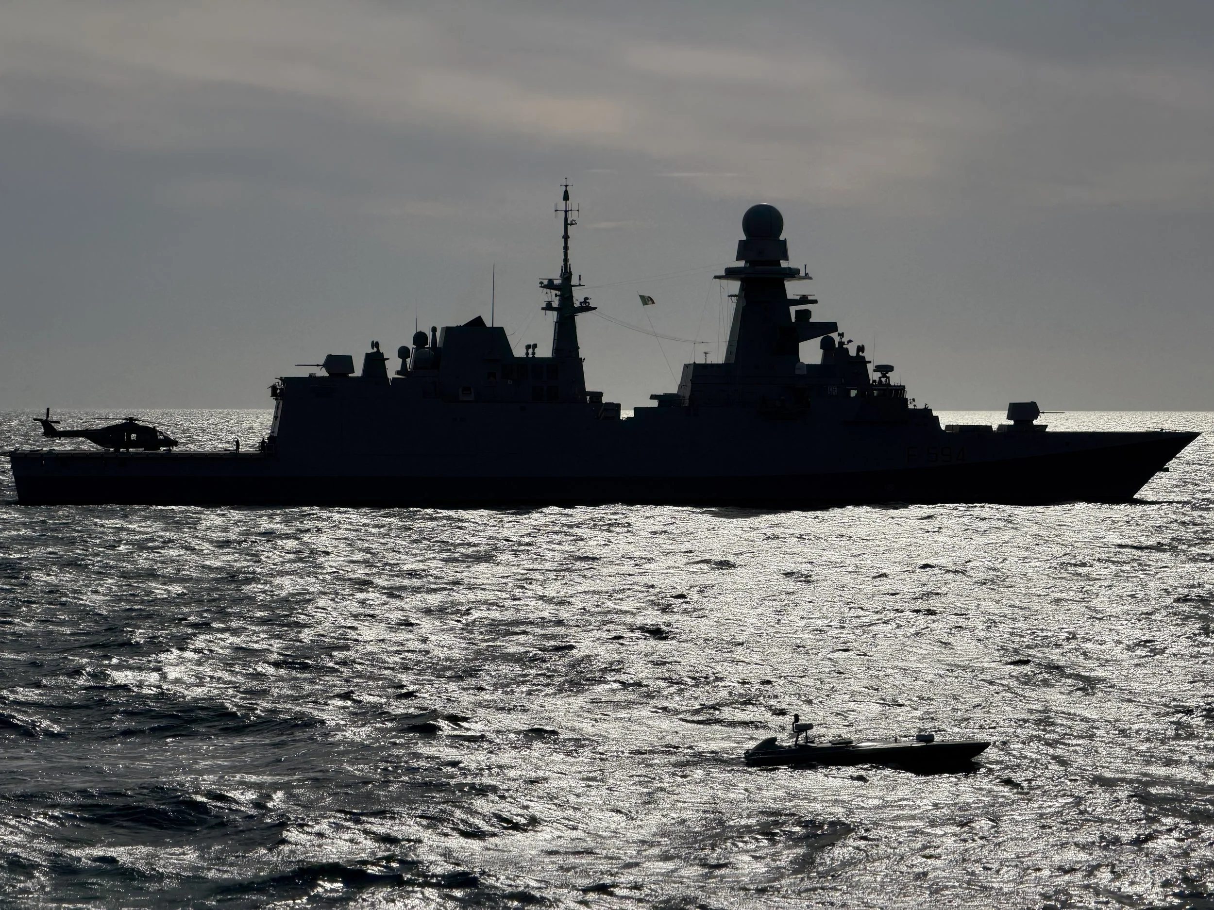 Silhouette of a large military ship with a helicopter flying nearby, on a body of water with the sun reflecting on the surface.