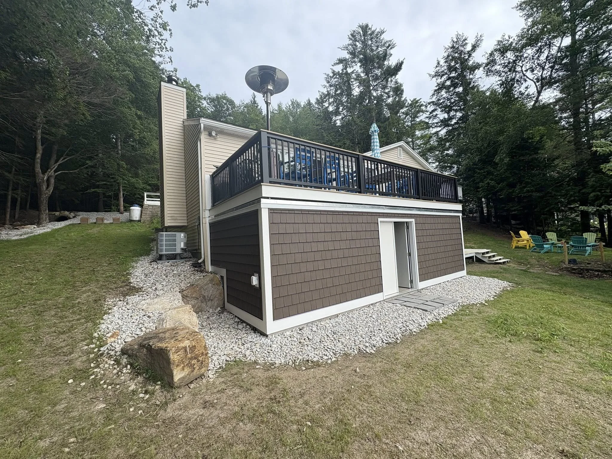 A two-story house with a deck on top, decorated with outdoor furniture and an umbrella, situated in a wooded outdoor area with Adirondack chairs in the background.